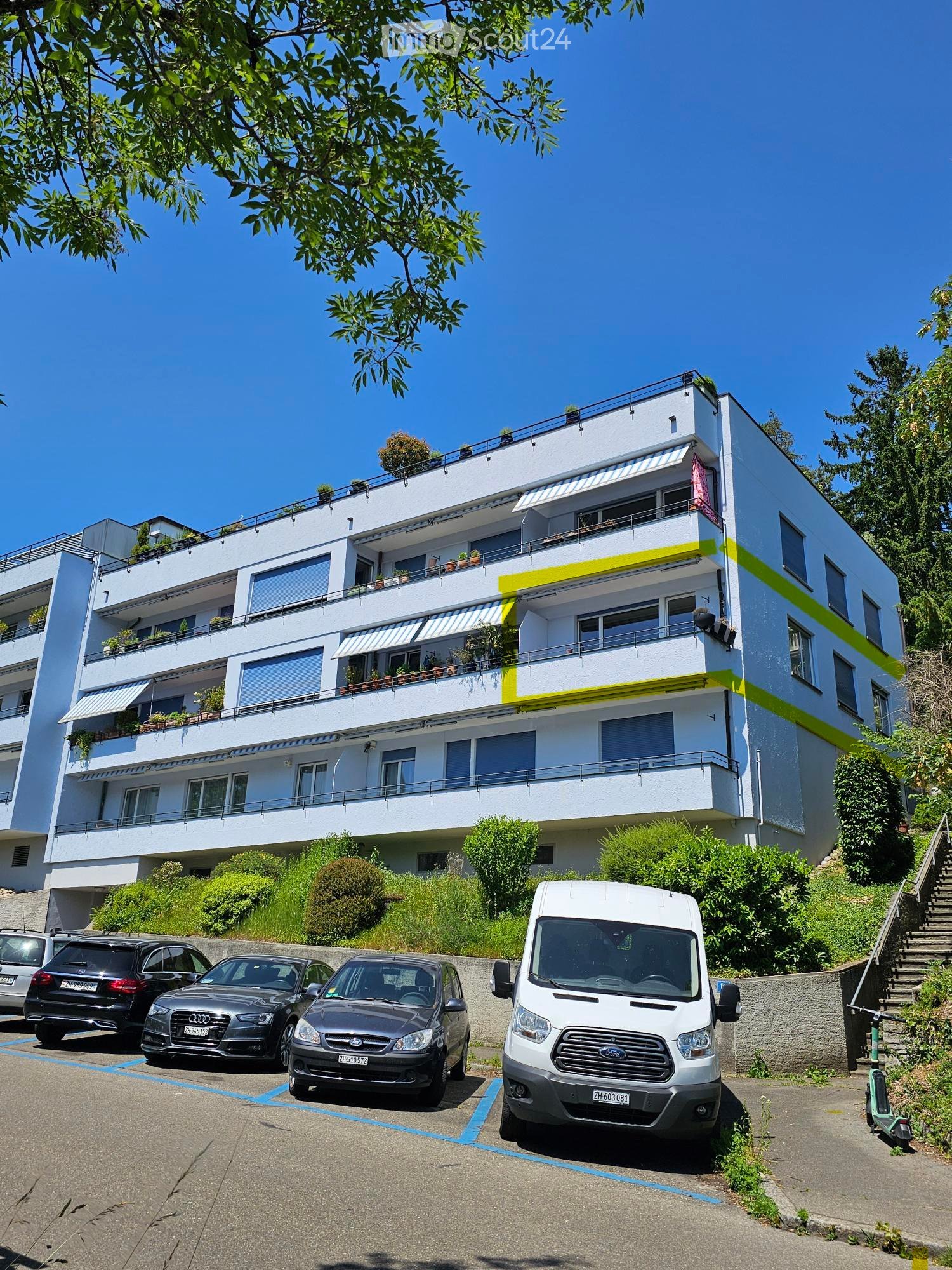 Modern multi-story apartment building with balconies and greenery, surrounded by parked cars and a staircase leading up.