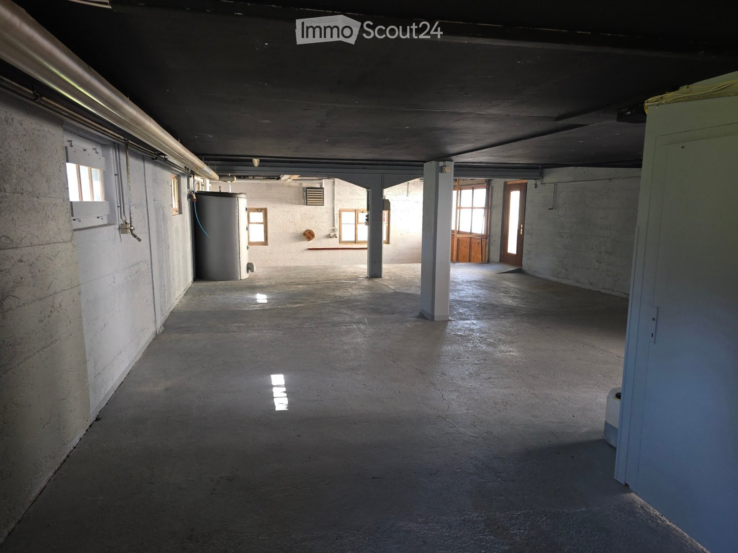 Empty concrete corridor with exposed pipes, columns, and small windows allowing natural light. Features a water tank and a closed door at the end.