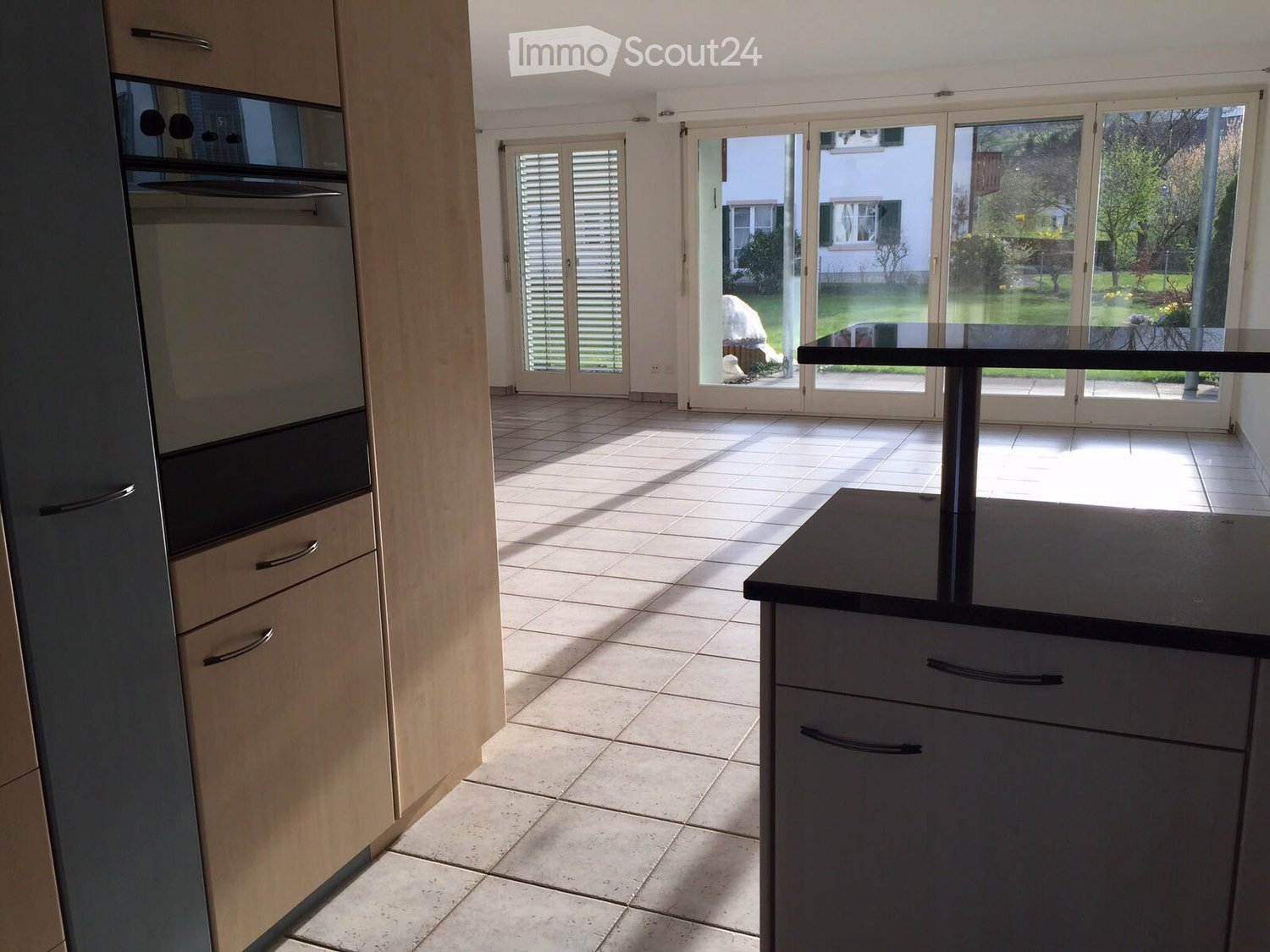 Modern kitchen with light wood cabinets, tiled floor, and large sliding glass doors leading to a garden.