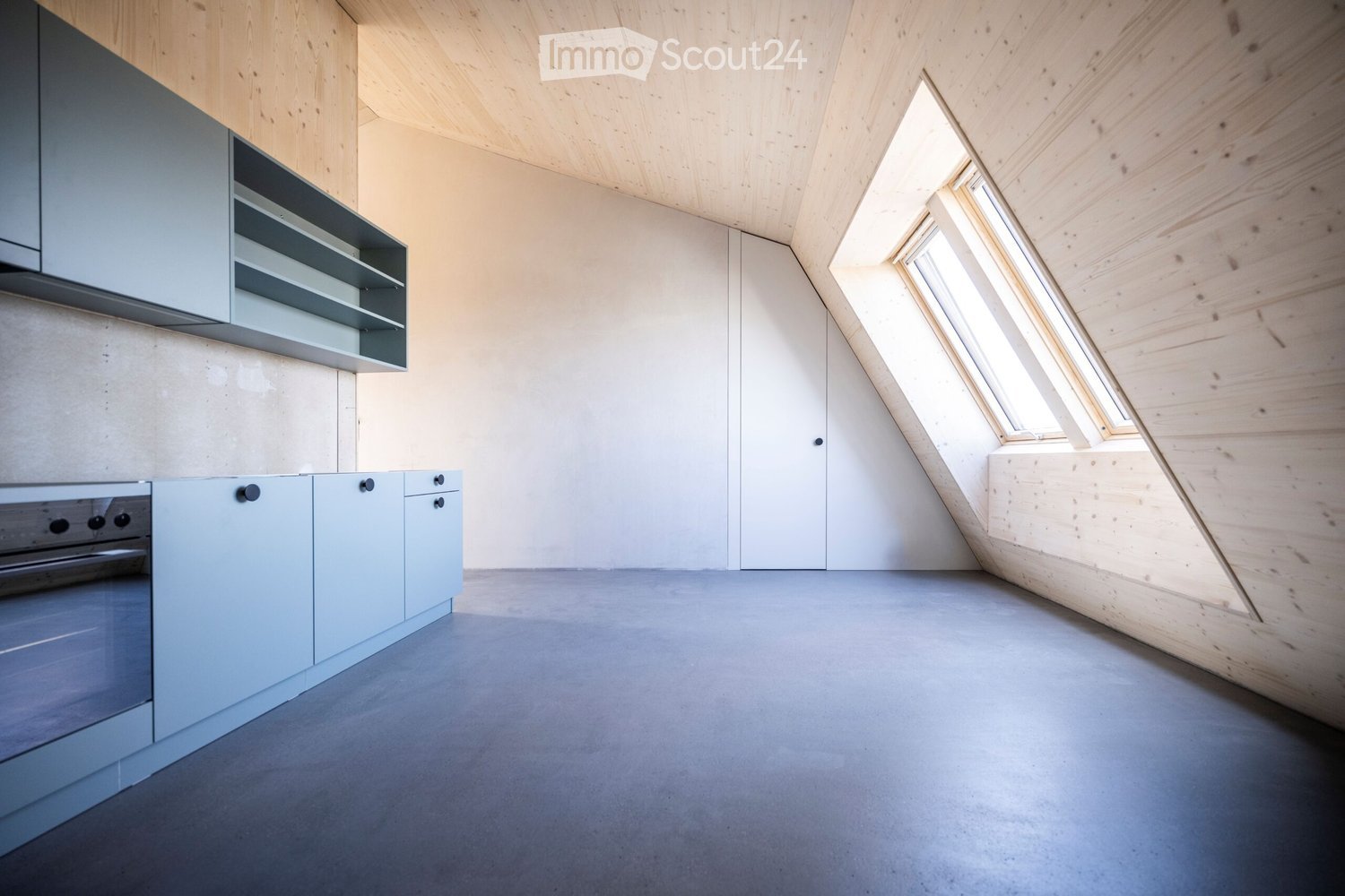 Modern kitchen with light grey cabinets, stainless steel oven, and built-in shelving, featuring a concrete floor and wooden ceiling with skylight windows.
