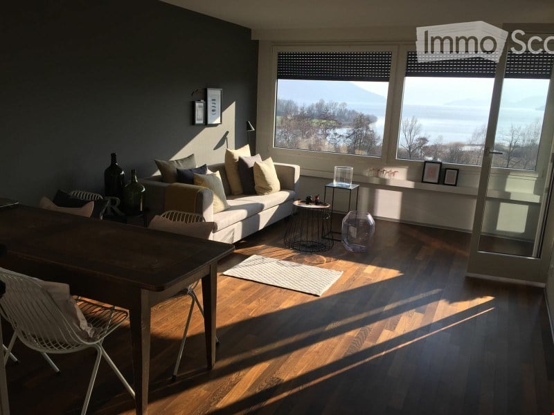 Bright living room with large windows offering a scenic view, featuring a white sofa with multiple cushions, a wooden coffee table, a dining table with chairs, and decorative vases.