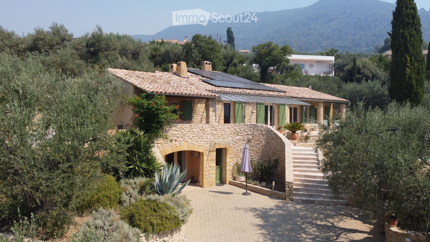 A two-story stone house with a tiled roof, green shutters, and a covered terrace area. The house is surrounded by lush greenery, including olive trees and various plants, with a scenic mountain view in the background.