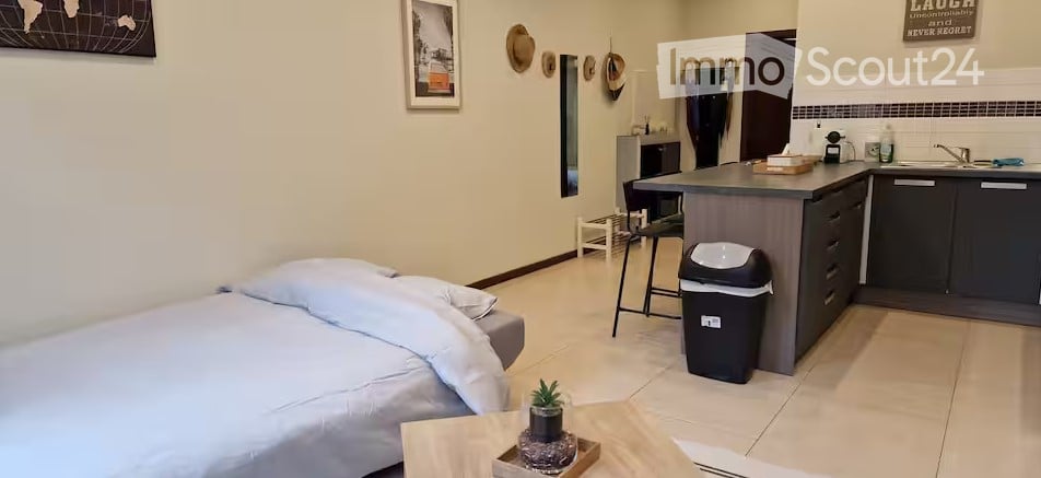 A compact studio apartment with a bed, small kitchen area, and a small table with a plant. The room features light-colored walls, a tiled floor, and a few decorative items including framed pictures and hats on the wall.