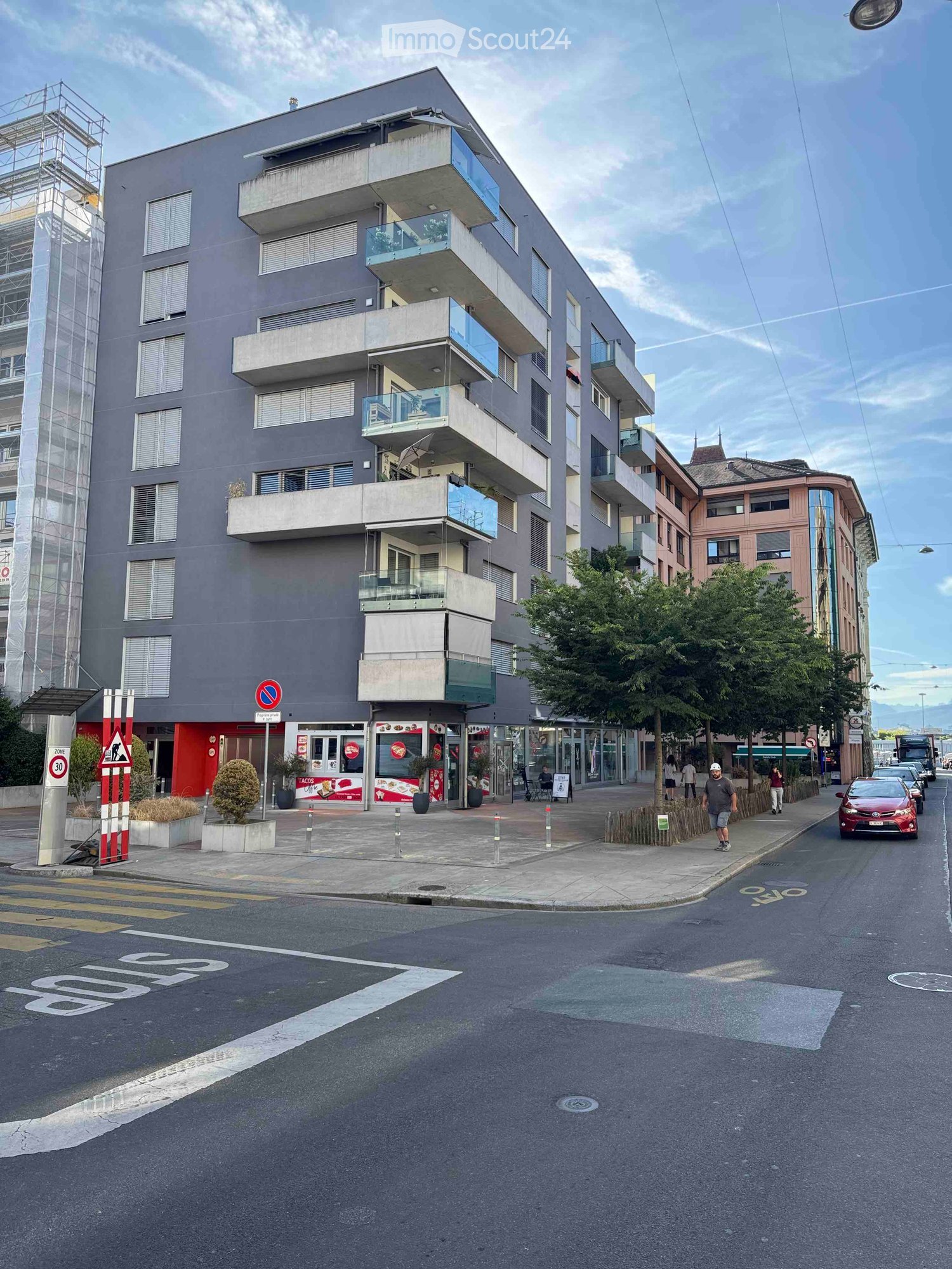 Modern multi-story building with balconies and large windows, located on a street corner with a small shop at the ground level.