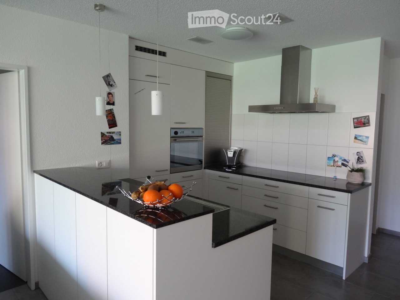 Modern kitchen with white cabinets, black countertops, and stainless steel appliances, including an oven and a coffee maker. A fruit bowl with oranges and bananas is placed on the counter.