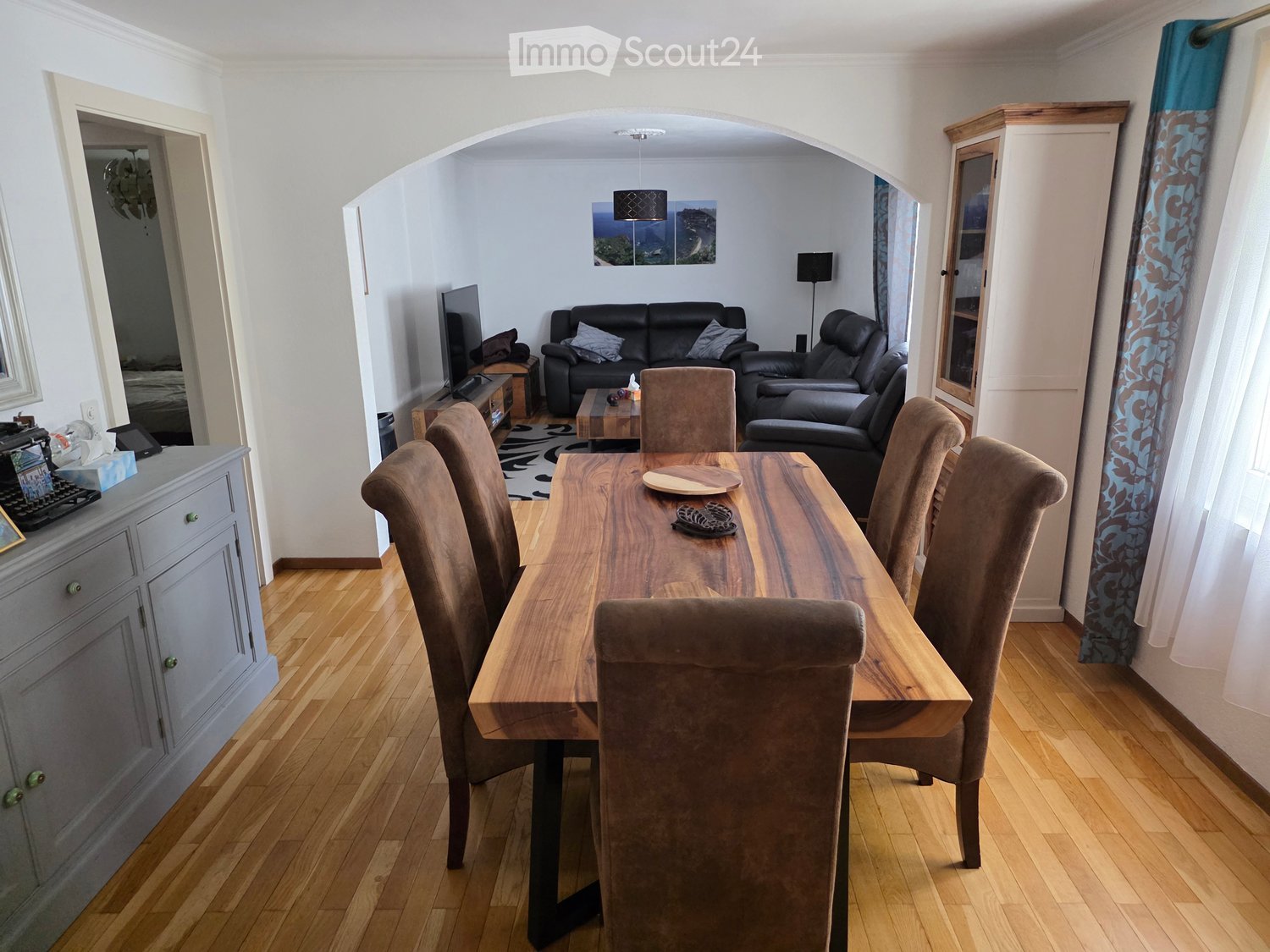 Dining area with a wooden dining table and chairs, a living area with a sofa and armchair, and a television. The room features wooden flooring and white walls.
