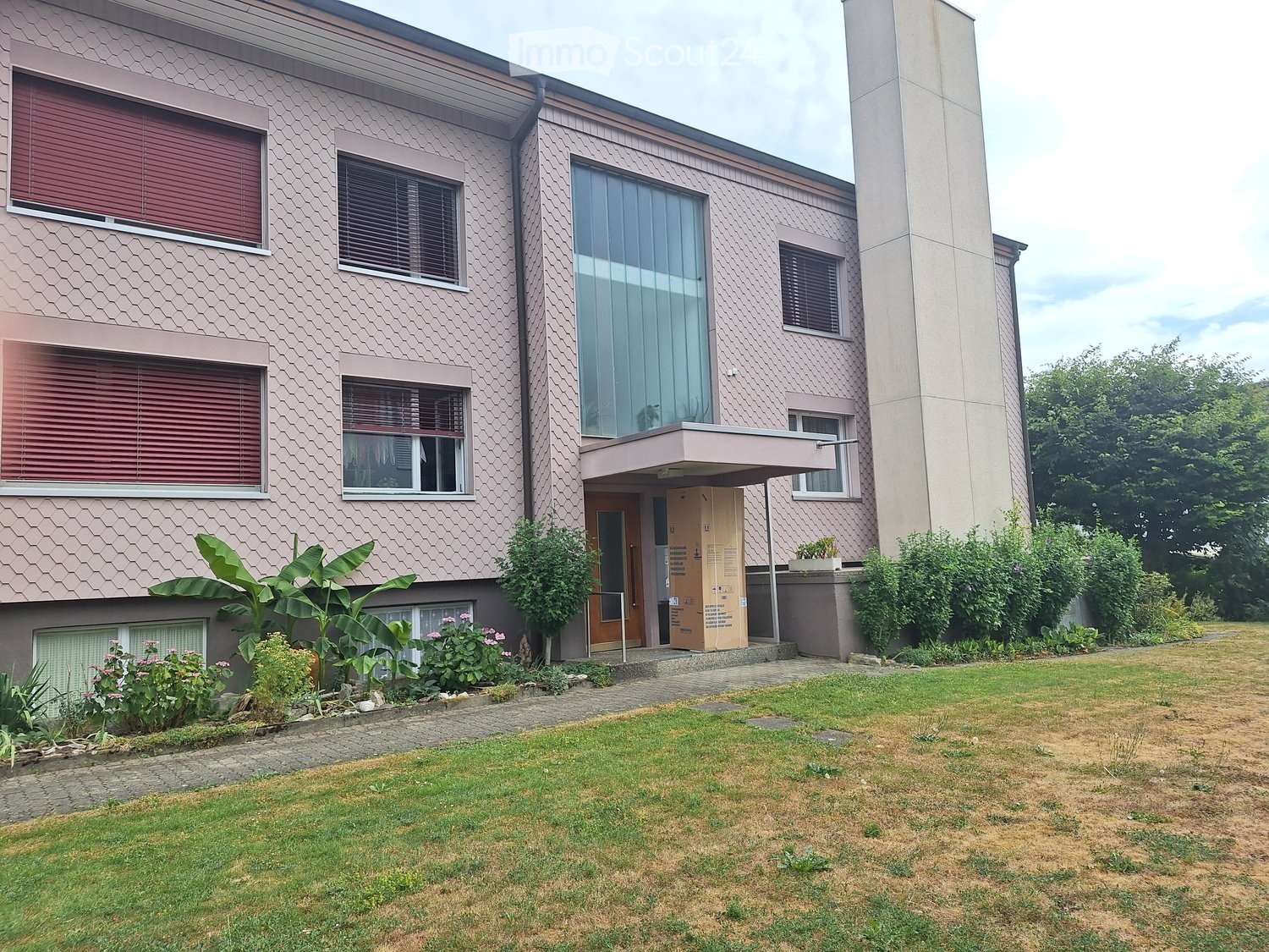 Two-story house with a textured facade, large windows, and a covered entrance. The front yard features a mix of green and brown grass, small plants, and a paved pathway leading to the entrance.