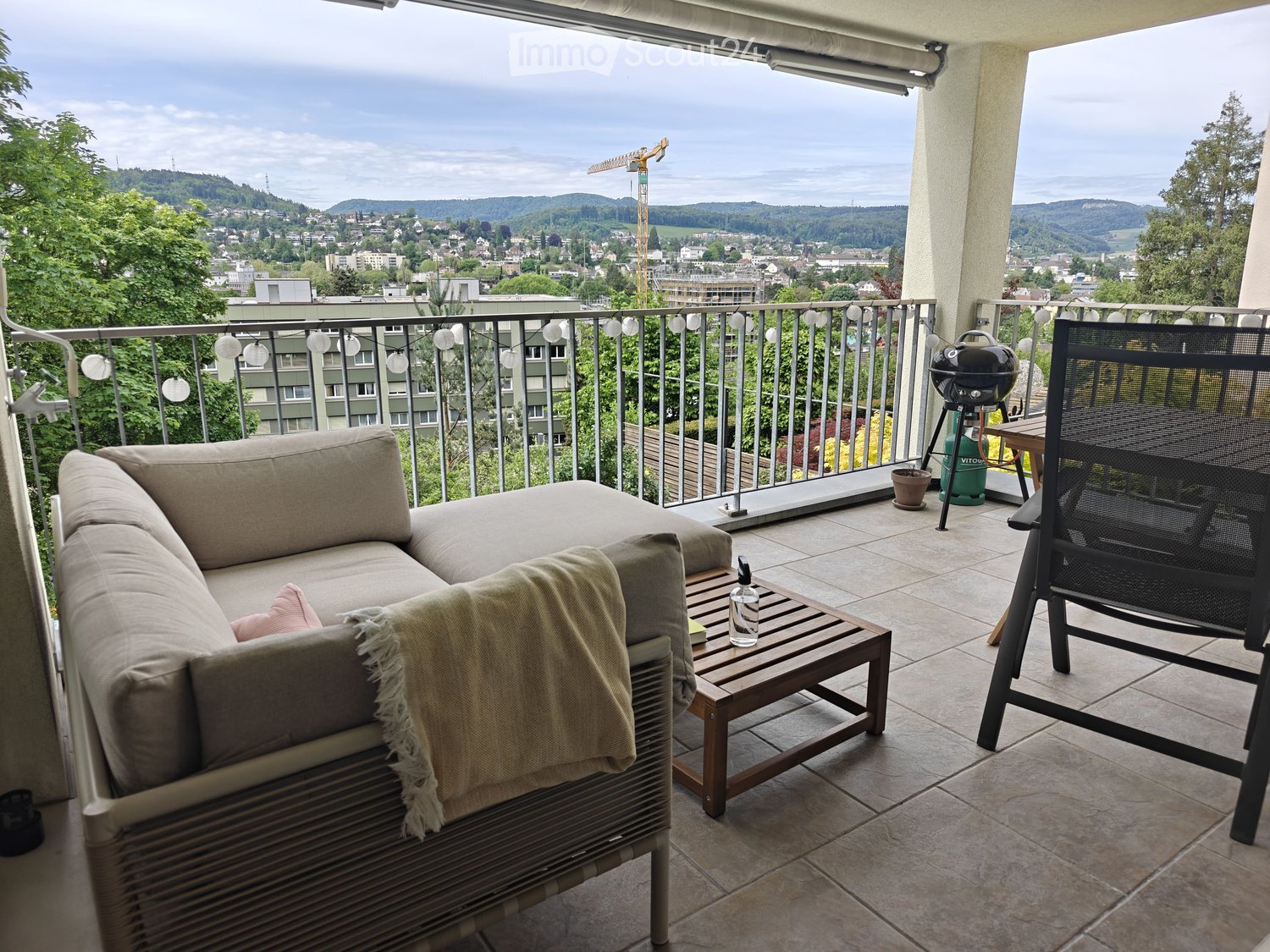 Balcony with a sofa, coffee table, chair, and grill, overlooking a cityscape with hills and trees.