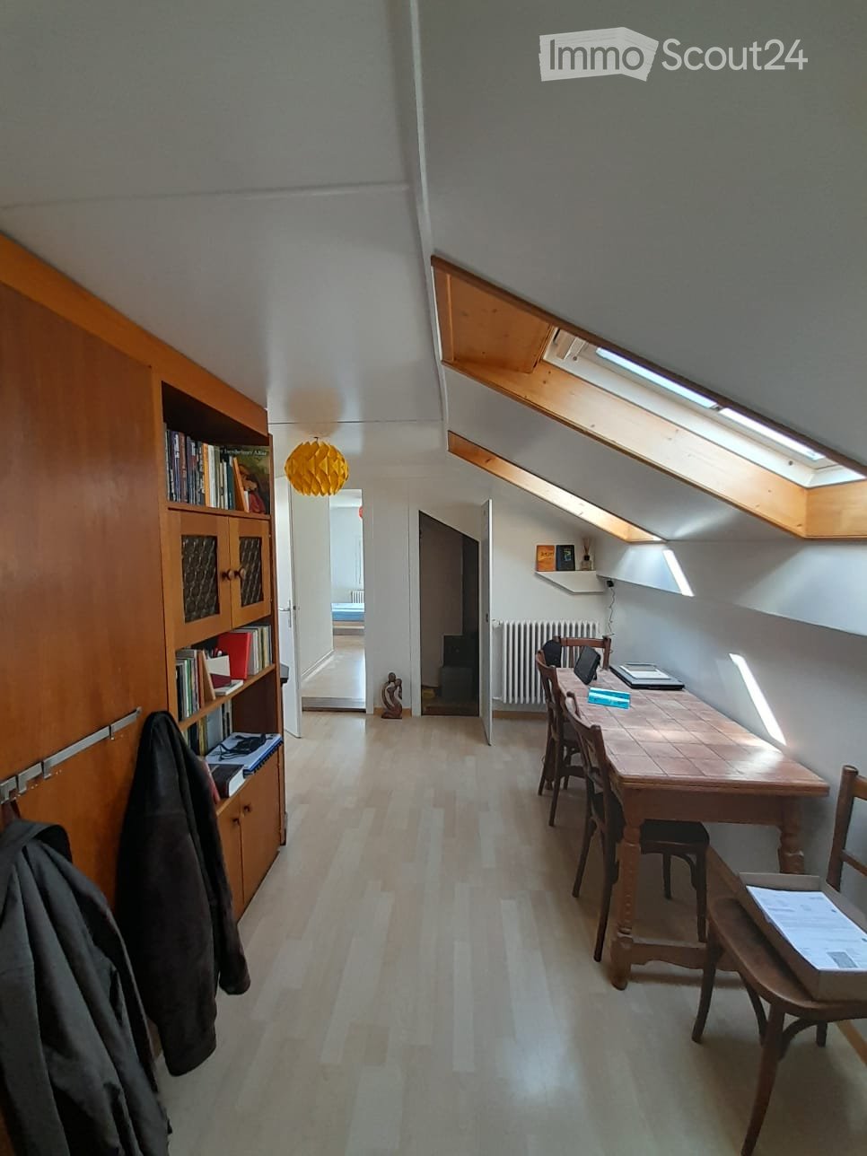 A narrow hallway with wooden flooring leads to a small, furnished room with a wooden table, chairs, a laptop, and a bookshelf. The room has slanted ceilings with skylights and a radiator.