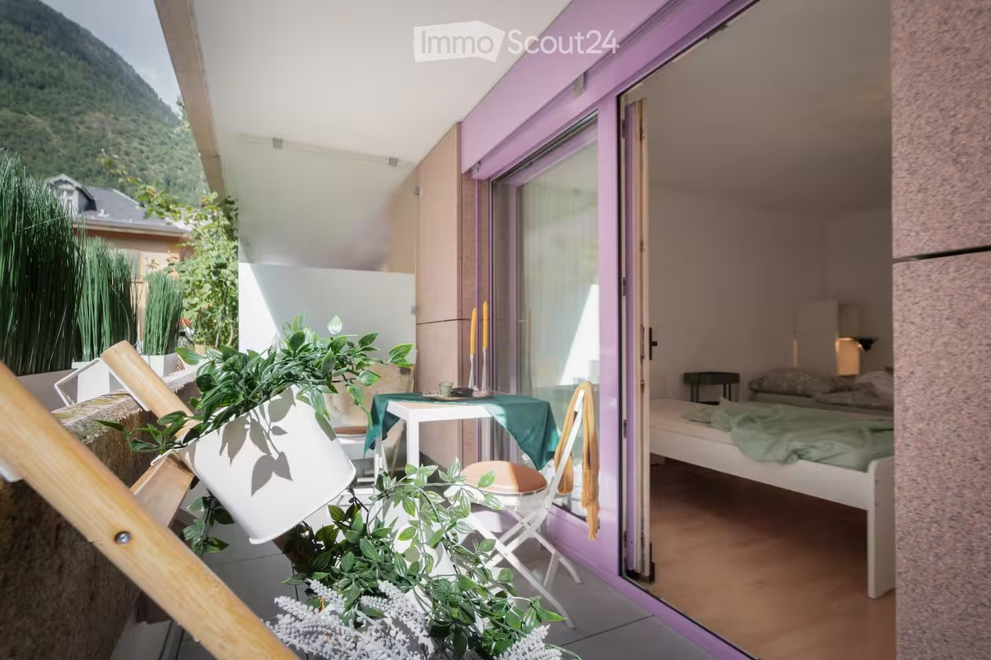 Balcony with a view of a mountain, featuring a small table with a green cloth, a chair, potted plants, and an open door leading to a bedroom with a bed and nightstand.