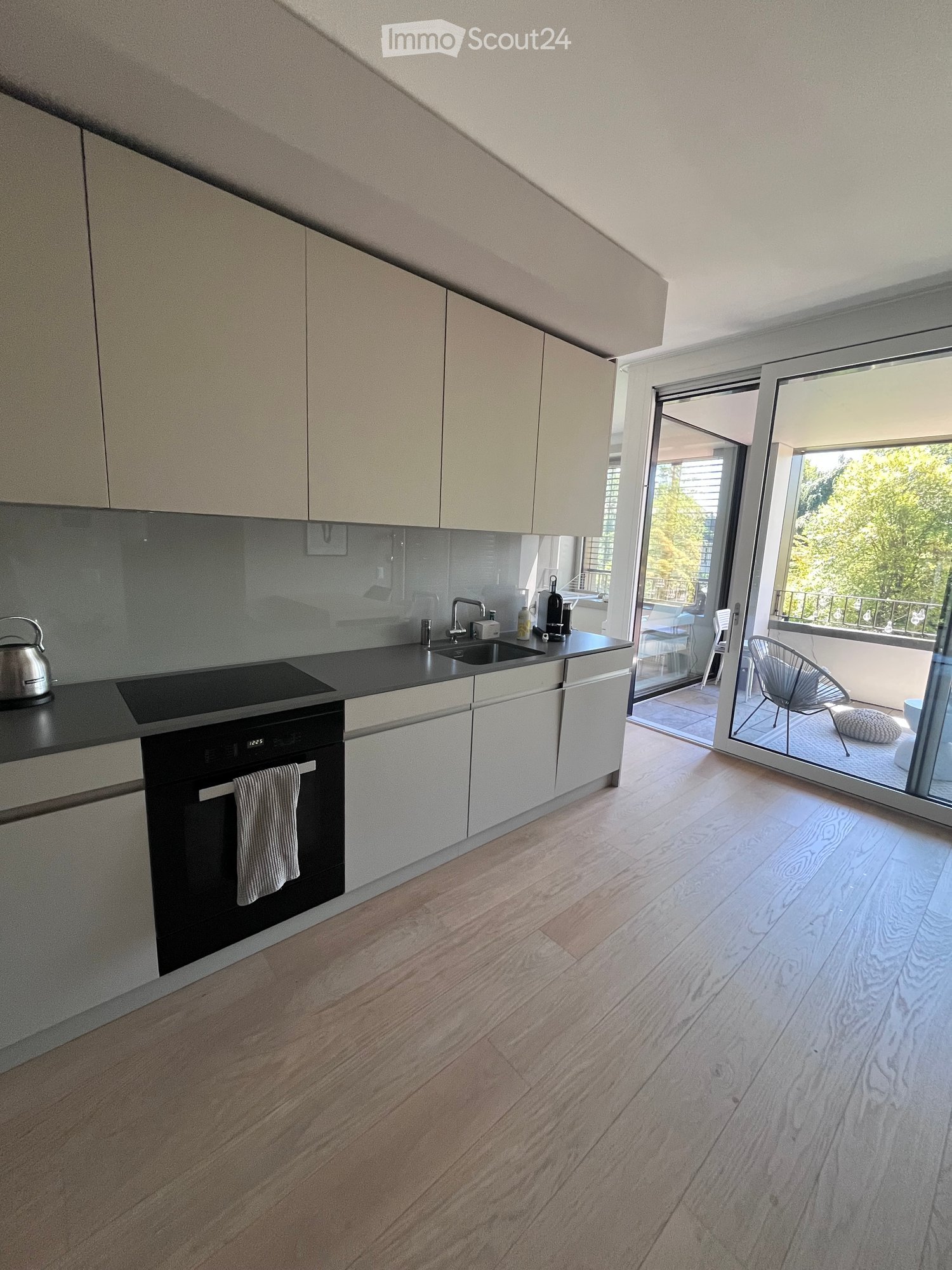 Modern kitchen with light-colored cabinets, wooden flooring, and a balcony view. Features an oven, sink, and various small appliances.