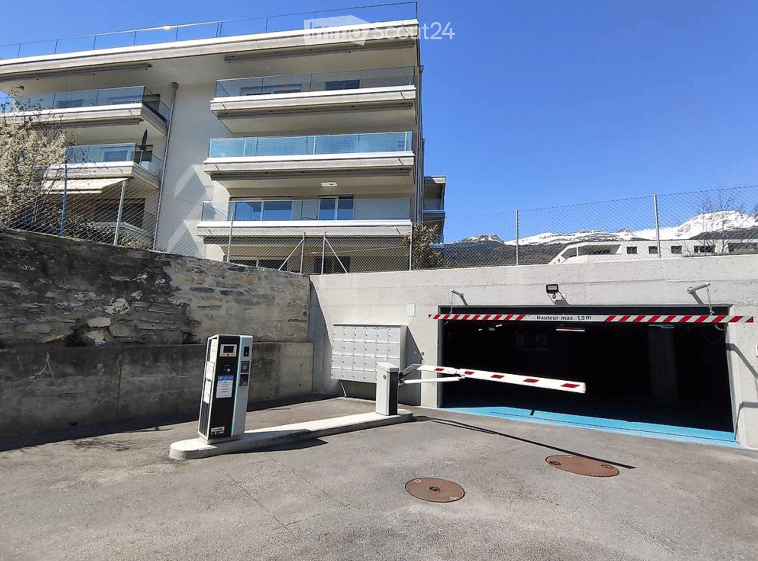 Modern multi-story building with balconies and a gated parking area. The entrance features a security gate and a parking barrier.