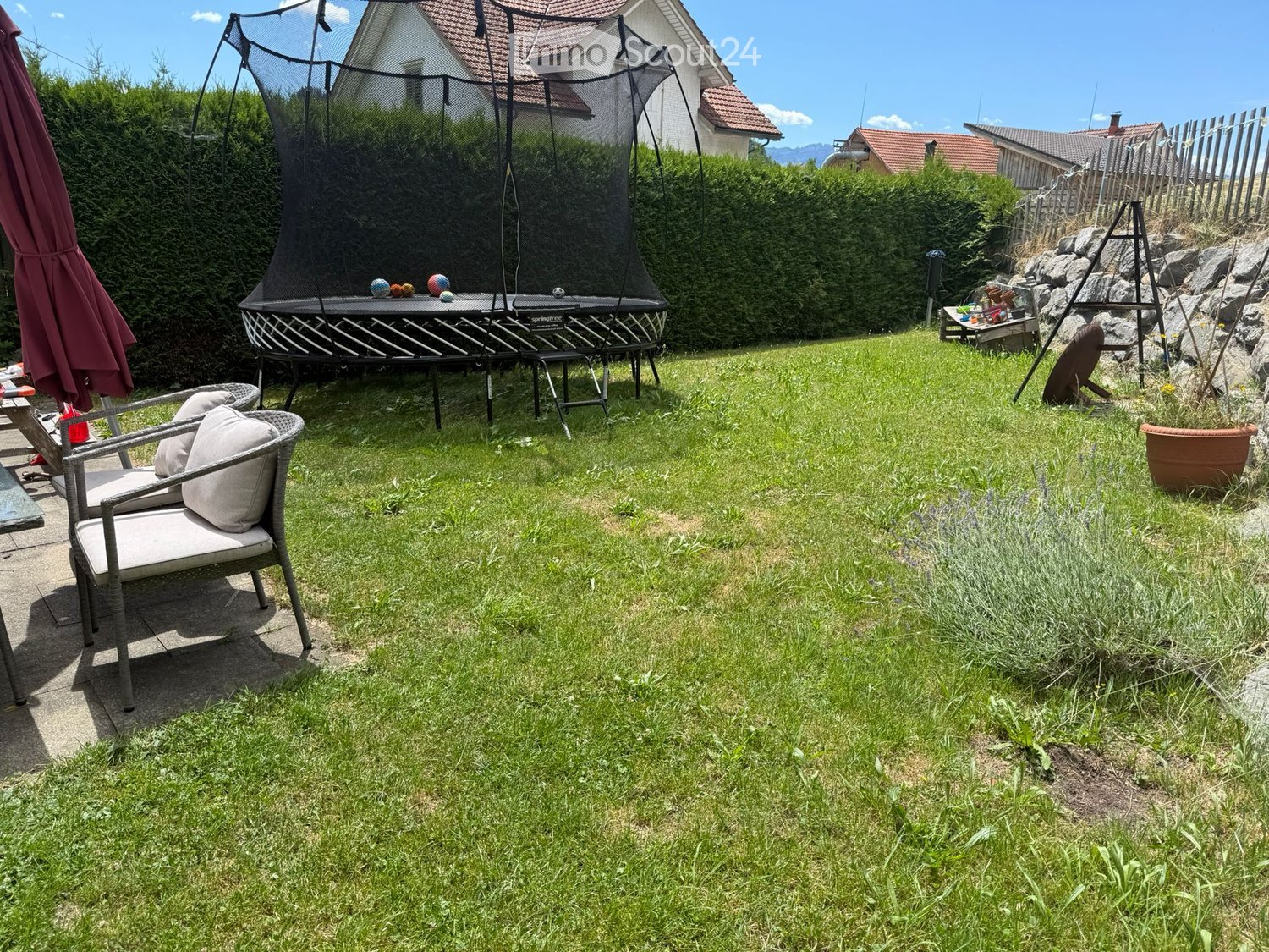 Lush green garden with a trampoline, a wicker chair with cushions, and a large umbrella on a patio.