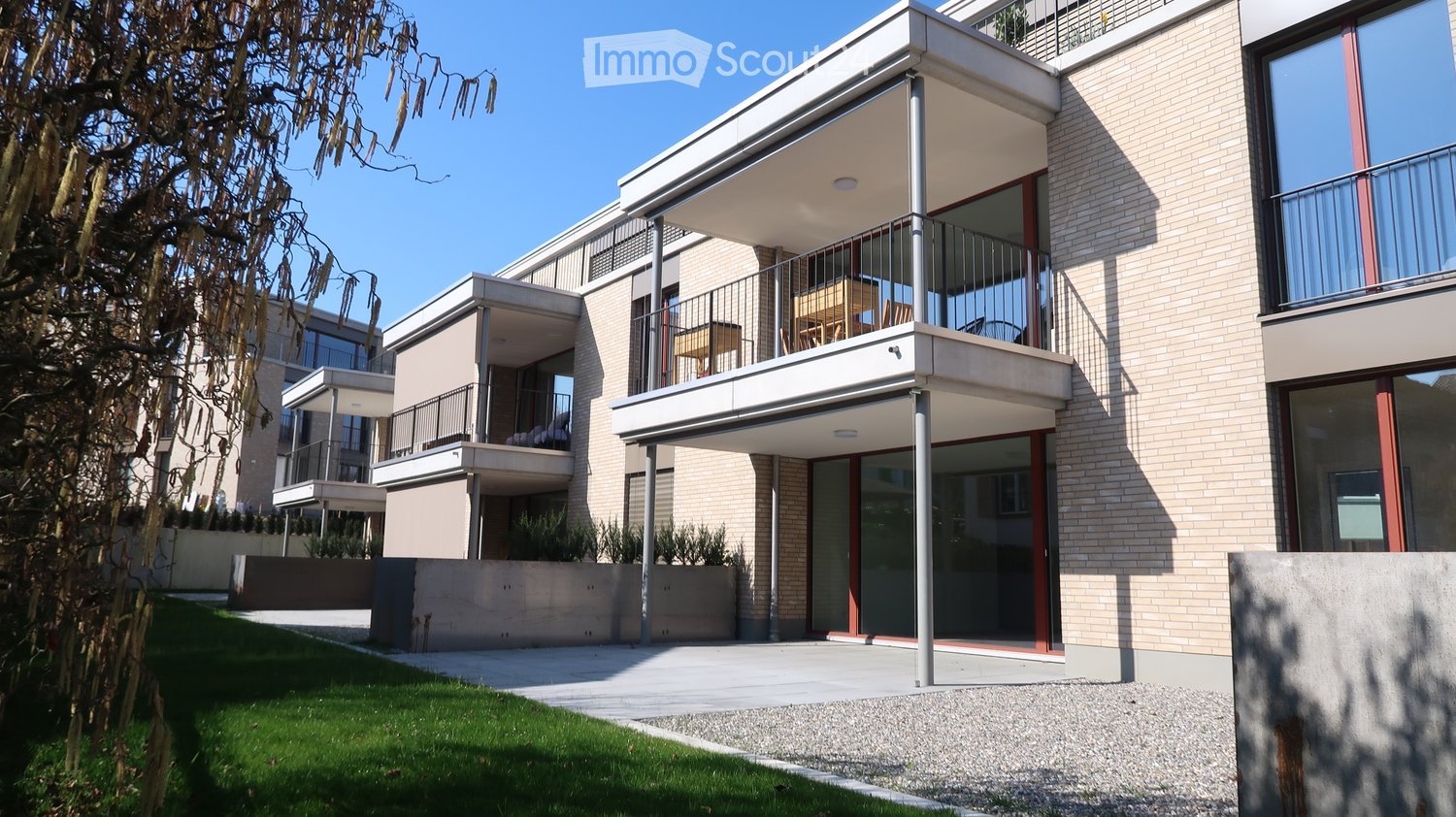 Modern multi-story building with balconies, large windows, and a clean, contemporary design. Features include a raised ground floor, a well-maintained lawn, and a paved pathway leading to the entrance.