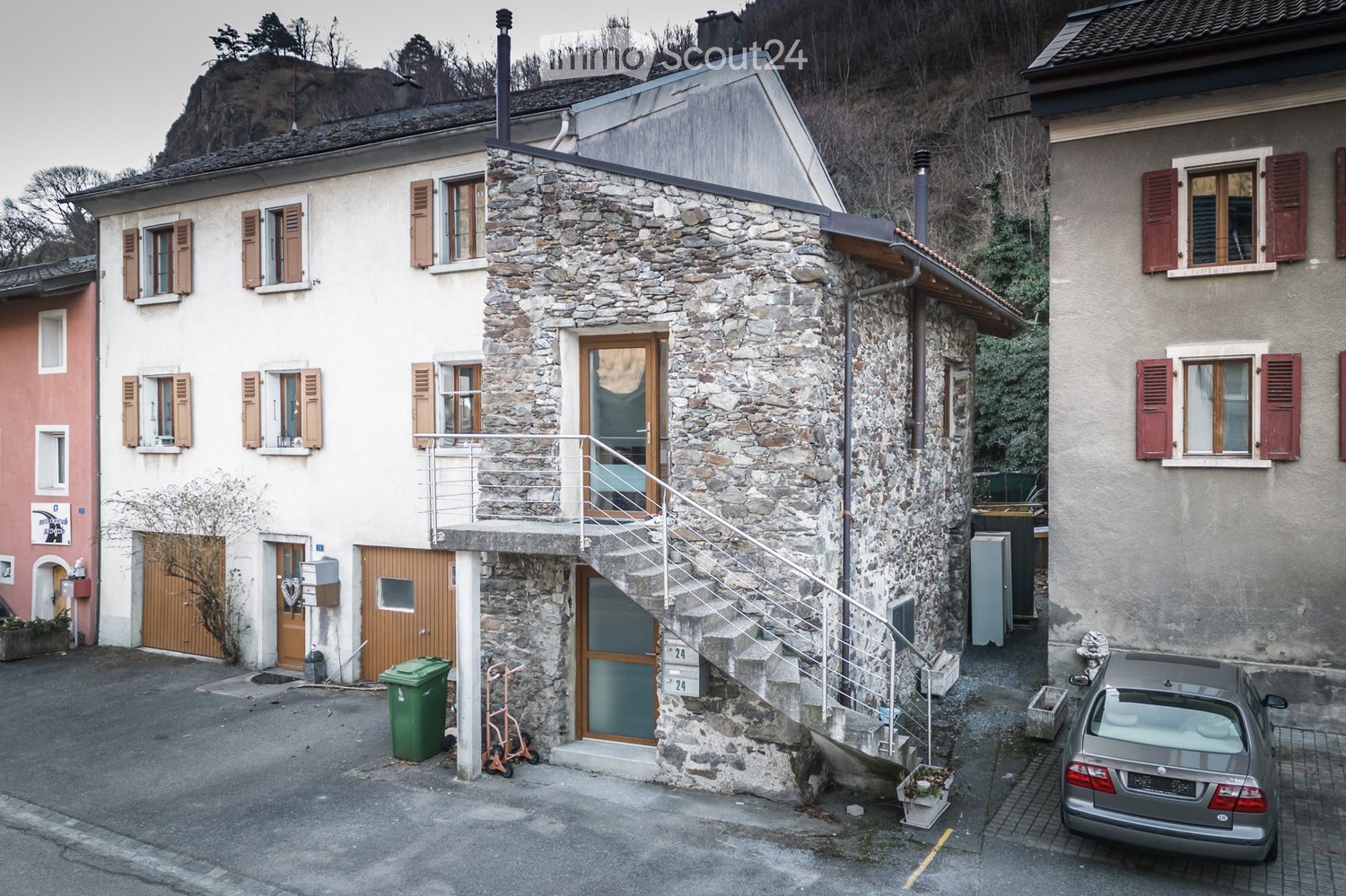 A two-story stone house with a balcony, wooden shutters, and a staircase leading to the entrance. A car is parked nearby, and there is a green trash bin and a small garden area.