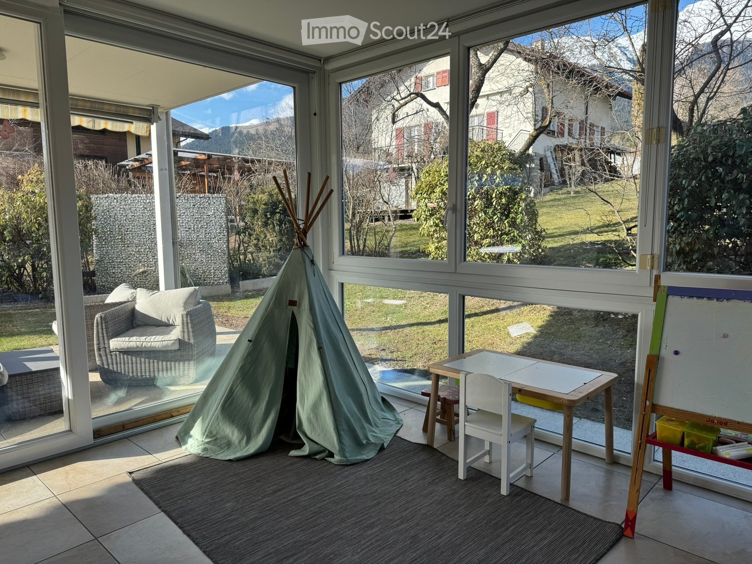 Bright room with large windows overlooking a garden, featuring a sofa, small table with chairs, a toy tent, and an easel with art supplies.