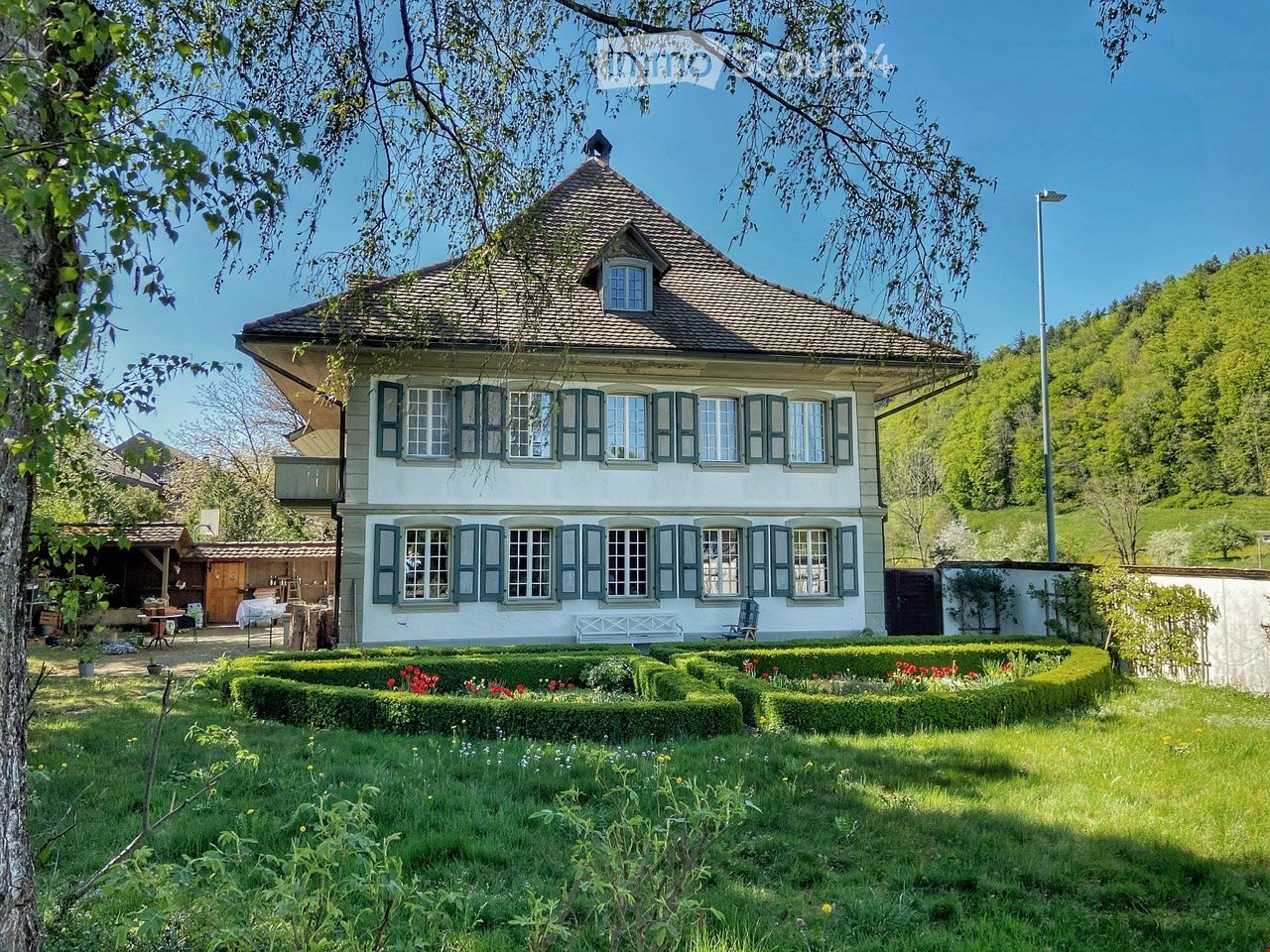 A two-story house with a traditional Swiss architectural style, featuring a pitched roof, multiple windows with shutters, and a well-maintained garden with neatly trimmed hedges and flower beds.
