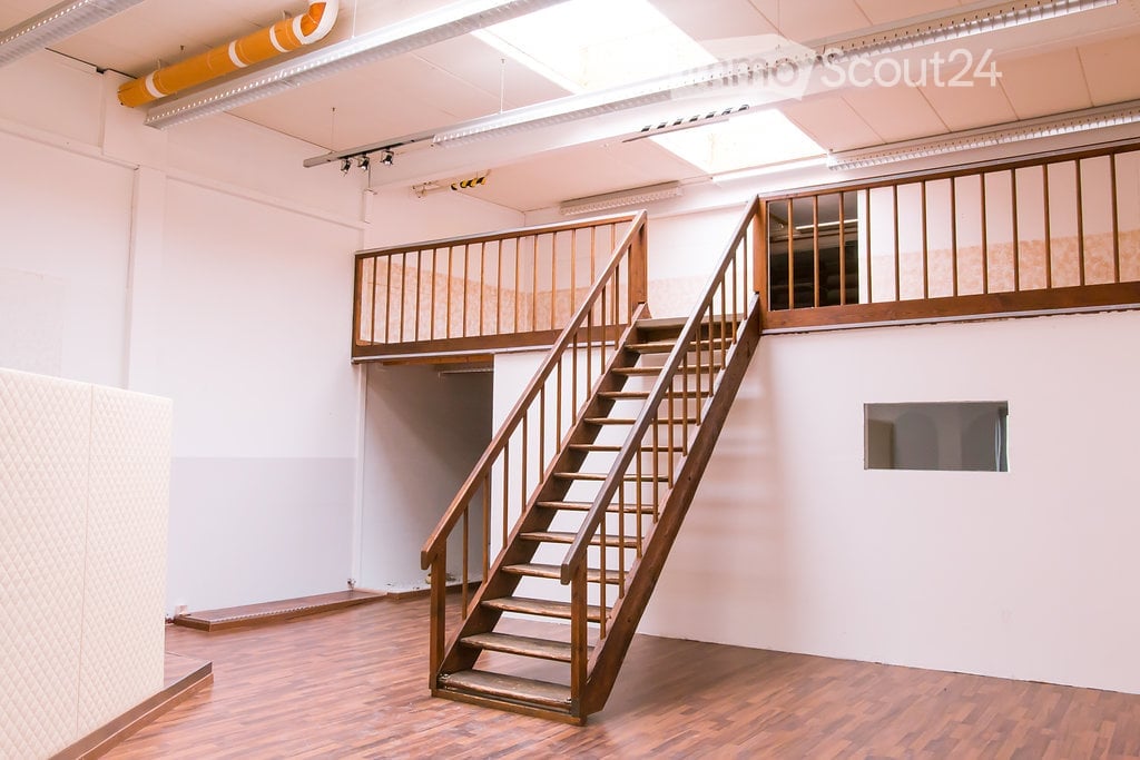 An empty room with wooden flooring, white walls, and a wooden staircase leading to an upper level with a wooden railing. The room has exposed ceiling beams and fluorescent lighting.