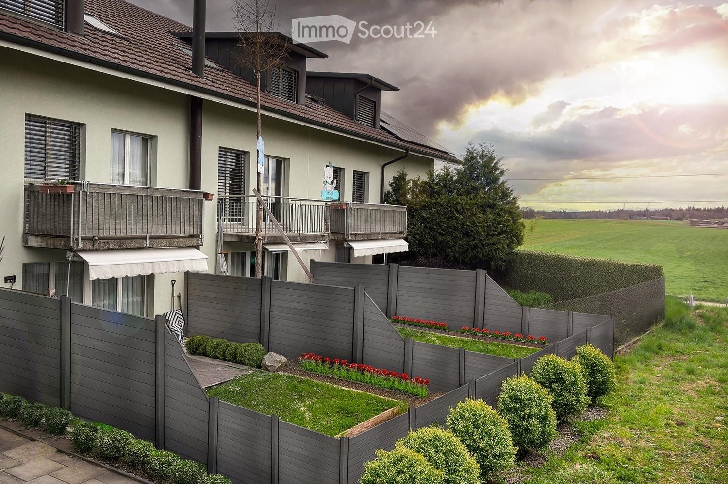 Two-story house with a tiled roof, multiple windows, and balconies. The surrounding area includes a well-maintained garden with a grey wooden fence, shrubs, and flower beds.