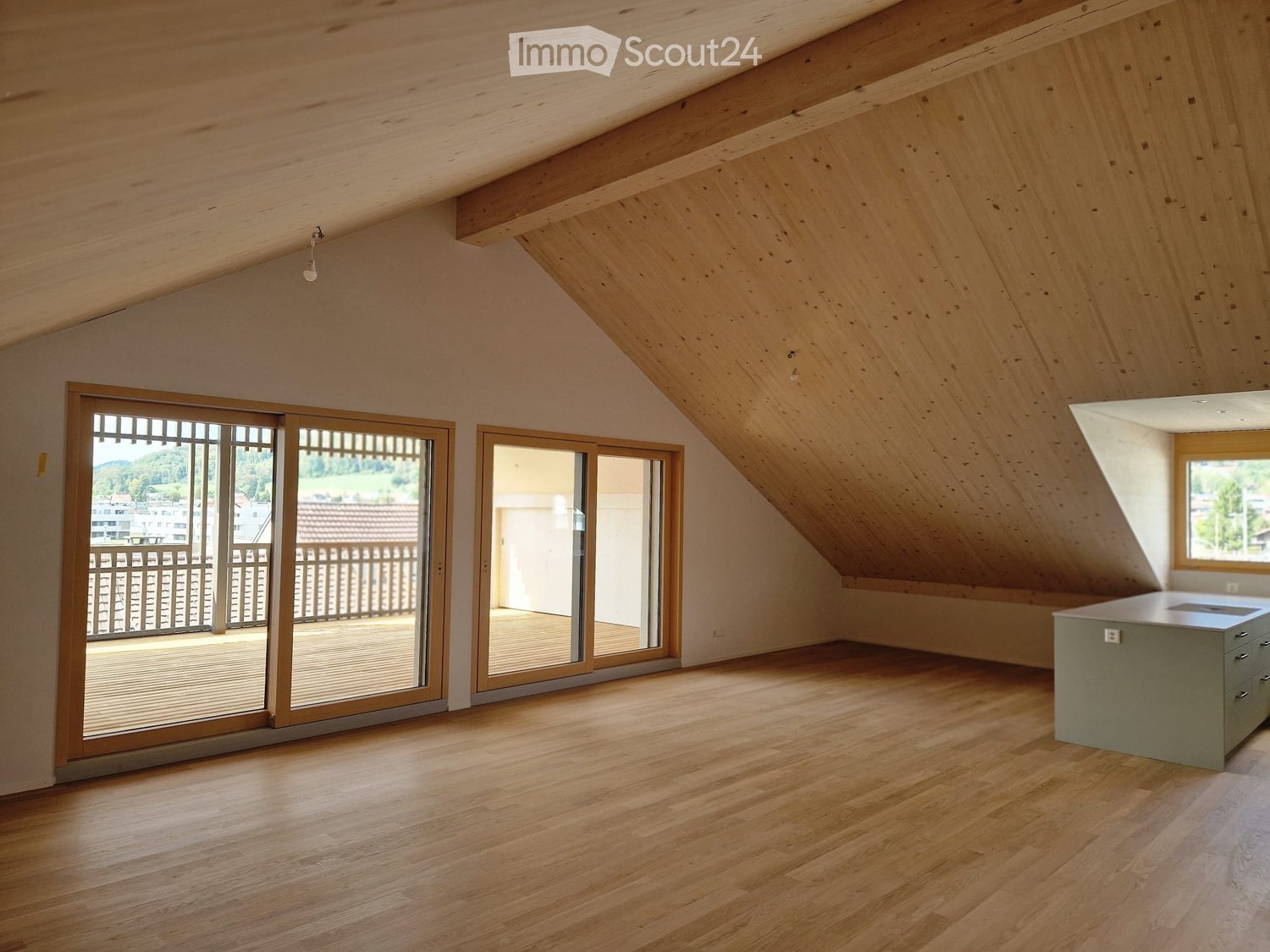 Bright, open attic space with wooden floors, exposed wooden beams, large windows, and sliding glass doors leading to a balcony.