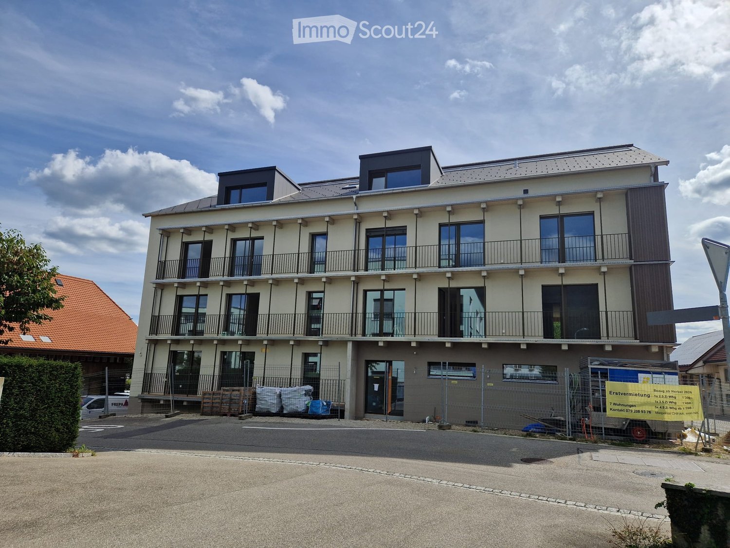 Modern multi-story residential building with balconies, large windows, and a contemporary design. The building has a mix of light-colored walls and dark accents, with a fenced-off area in front.