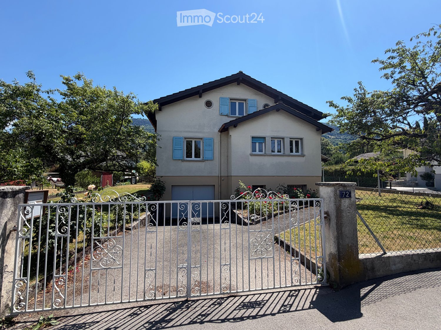 Two-story house with light-colored walls, blue shutters, and a garage. The property is surrounded by a white metal gate, greenery, and trees.