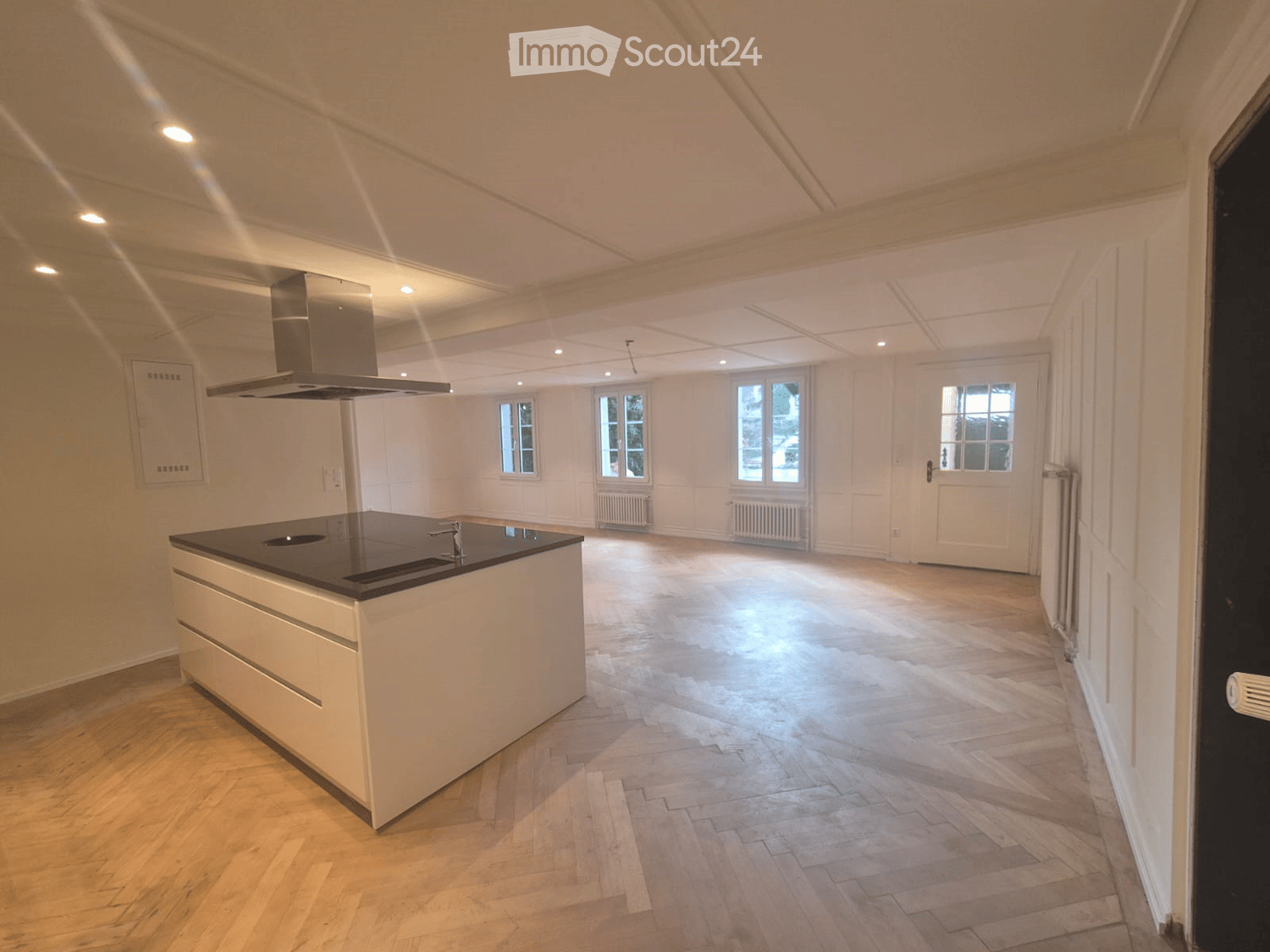 Open kitchen with island, wooden parquet floor, white walls, ceiling with recessed lighting, windows, kitchen hood, central island with sink and stove