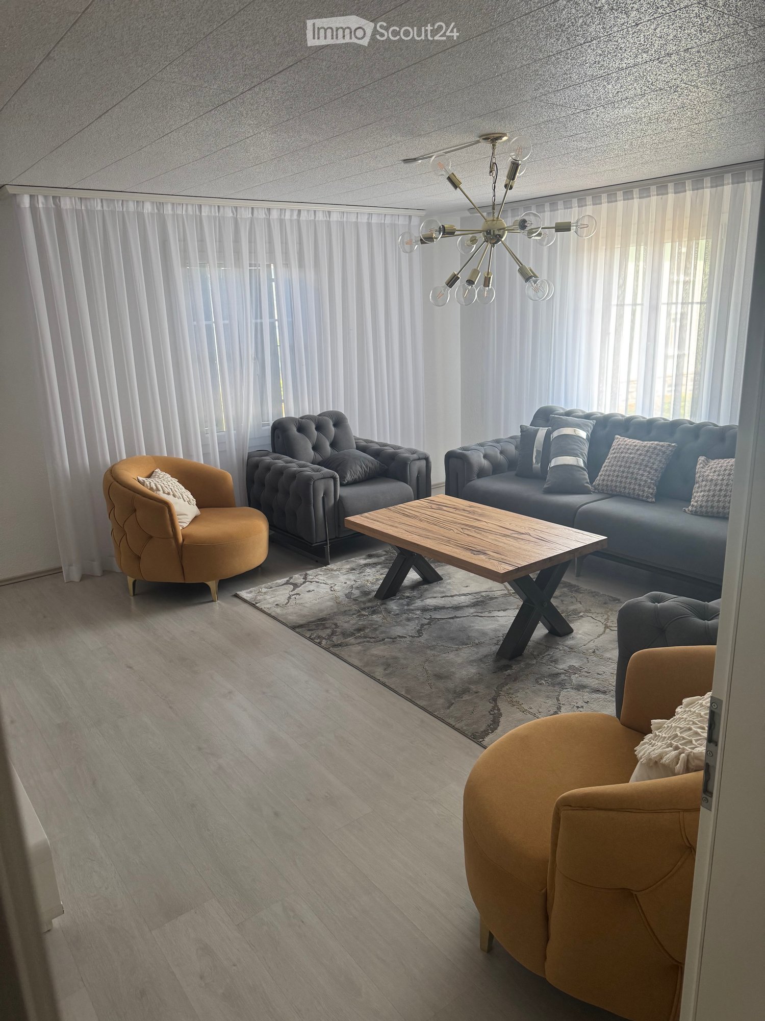 Modern living room with grey sofa, wooden coffee table, armchairs, chandelier, and large windows with white curtains.