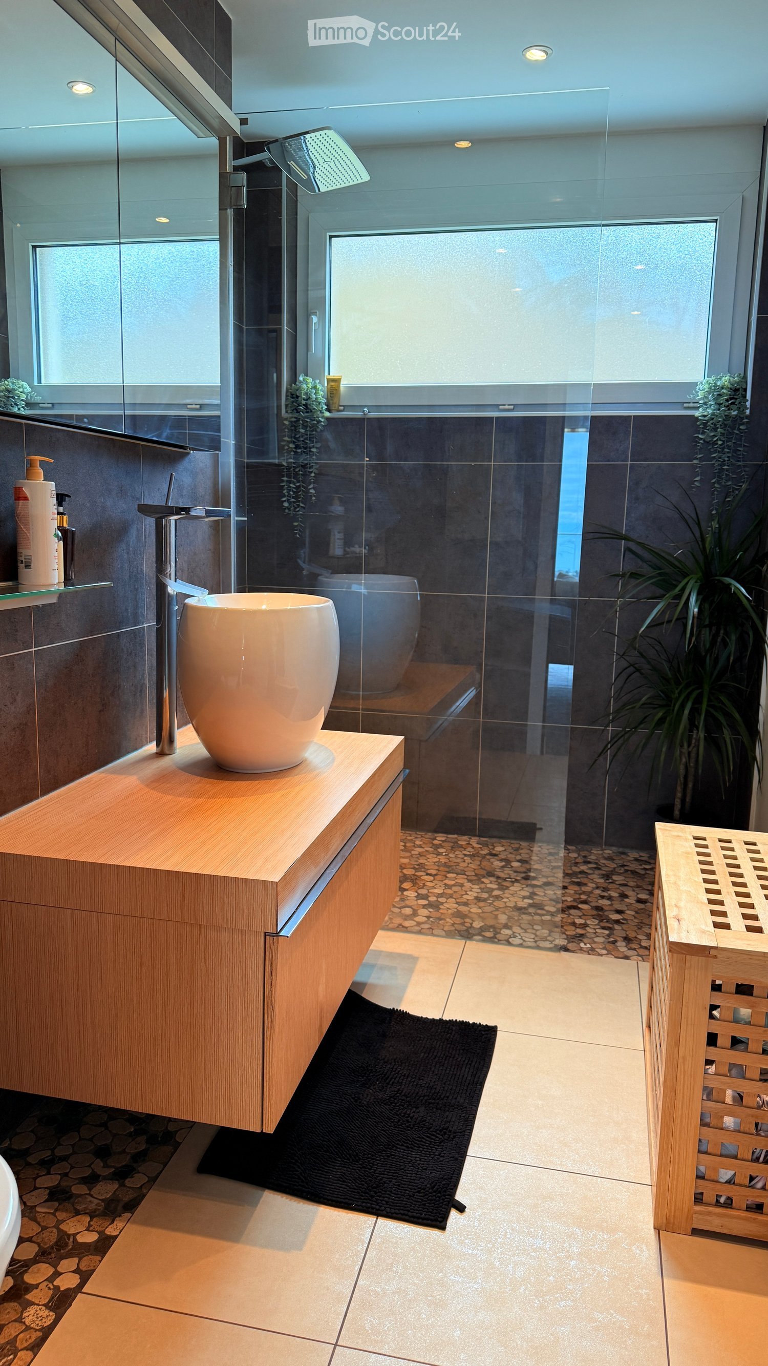 Modern bathroom with a wooden vanity, large sink, glass-enclosed shower, and a black bath mat.