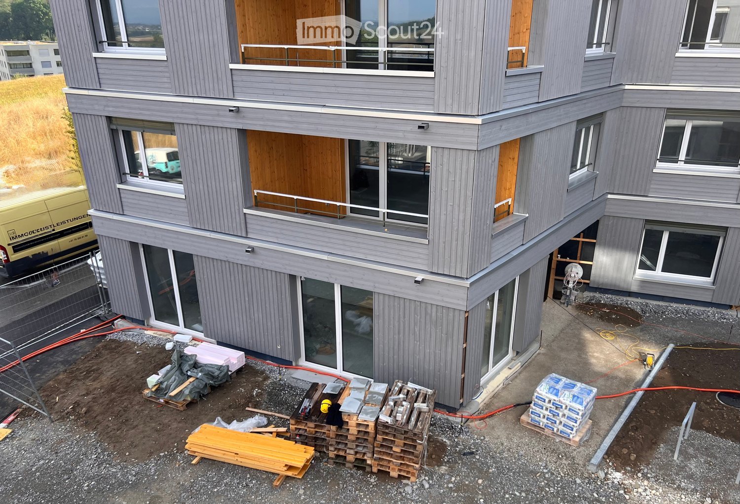 Modern two-story building with grey siding, large windows, and balconies. Construction materials and tools are visible on the ground.