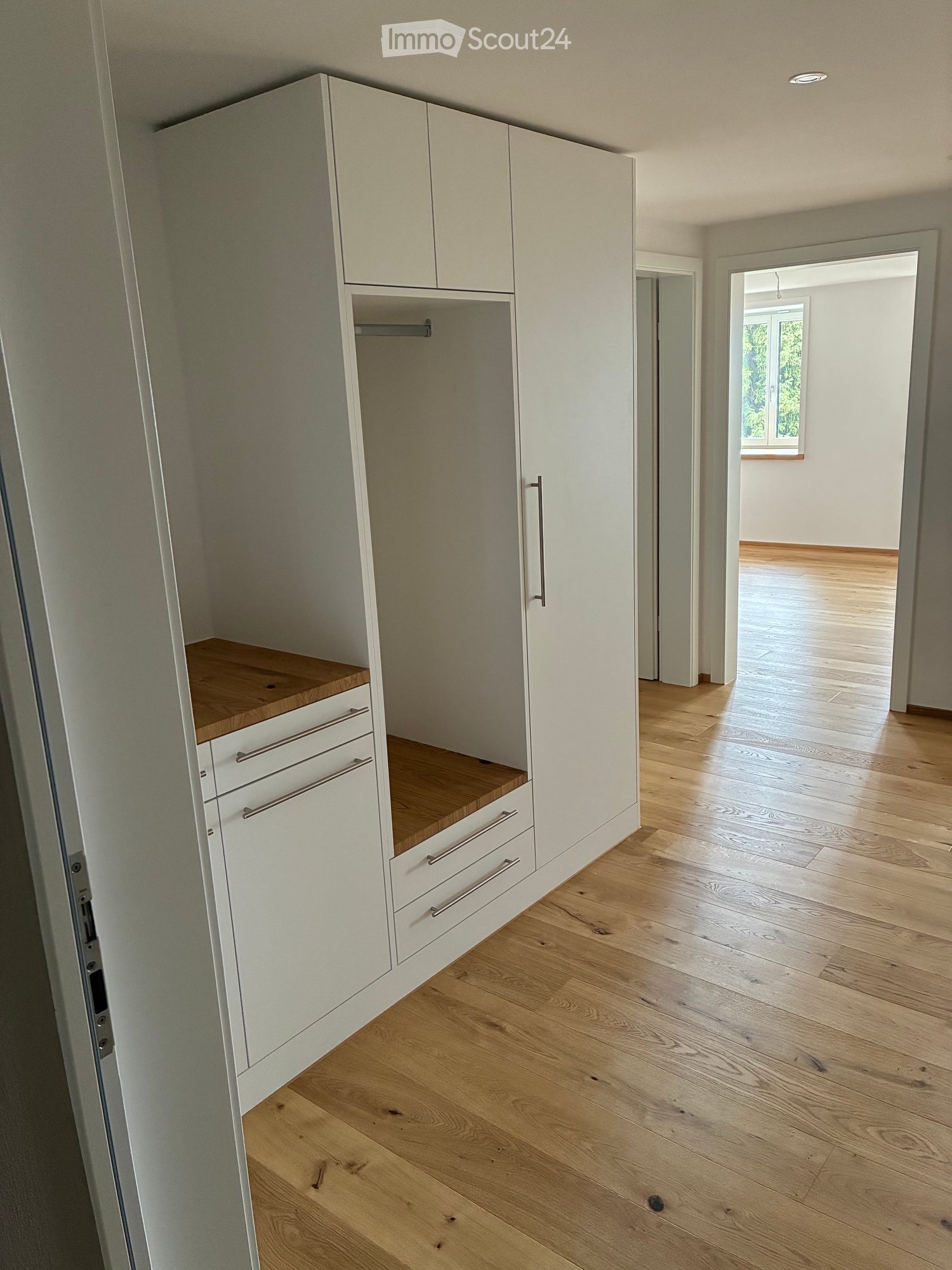 Empty room with light wooden flooring and white built-in wardrobe with drawers and shelves. The room is well-lit with natural light coming from an adjacent space.