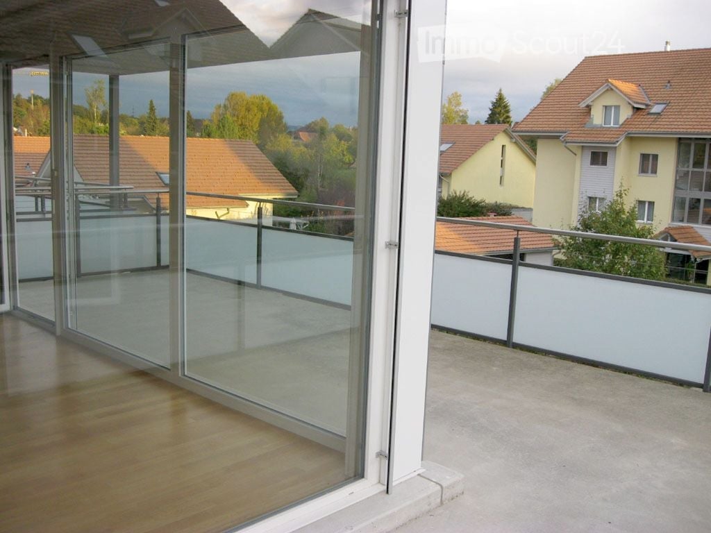Modern balcony with large glass sliding doors, wooden floor, white railing, and scenic view of neighboring houses and trees.