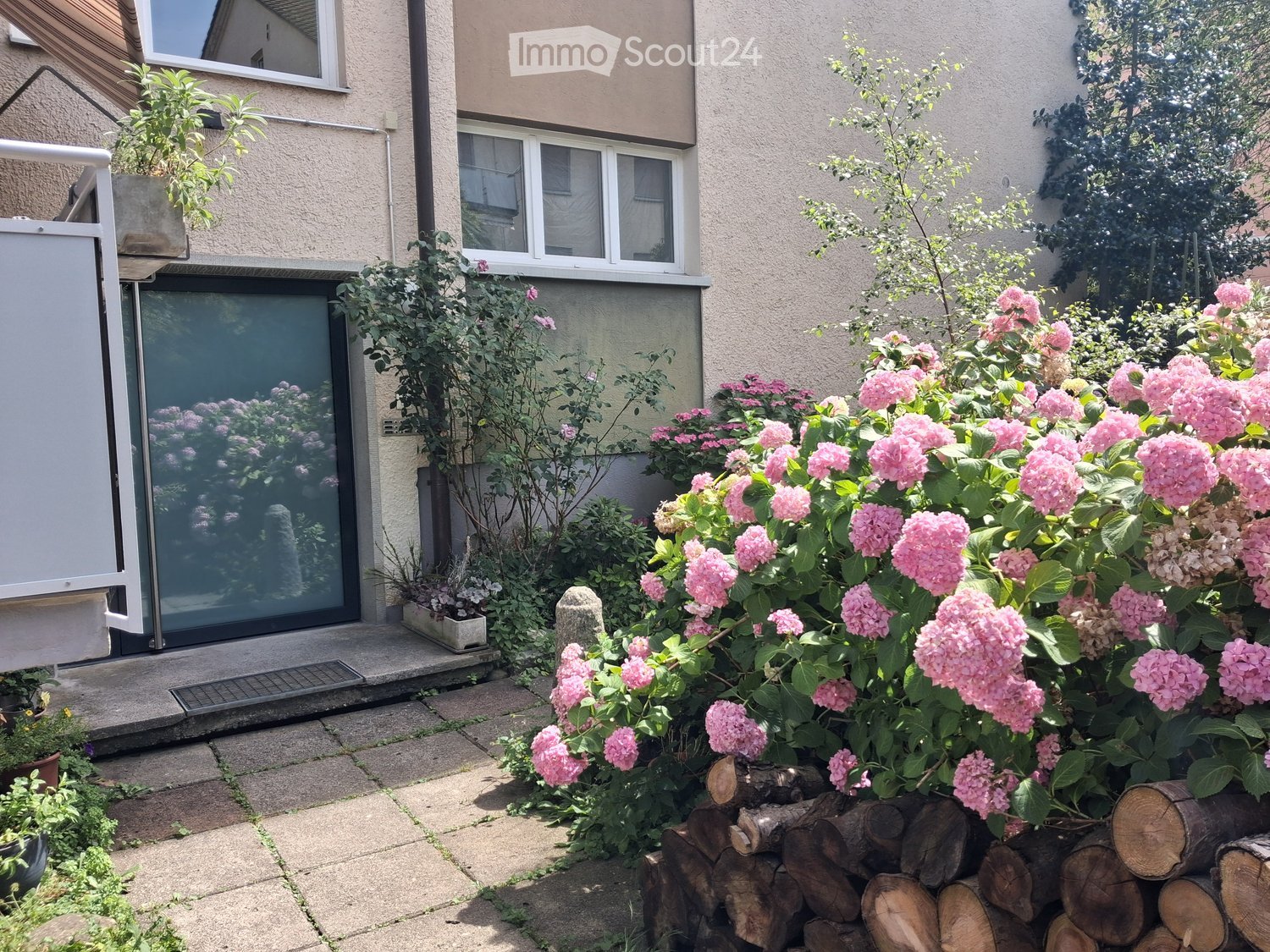 Garden area with pink flowers, stacked logs, and a paved patio next to a house entrance.