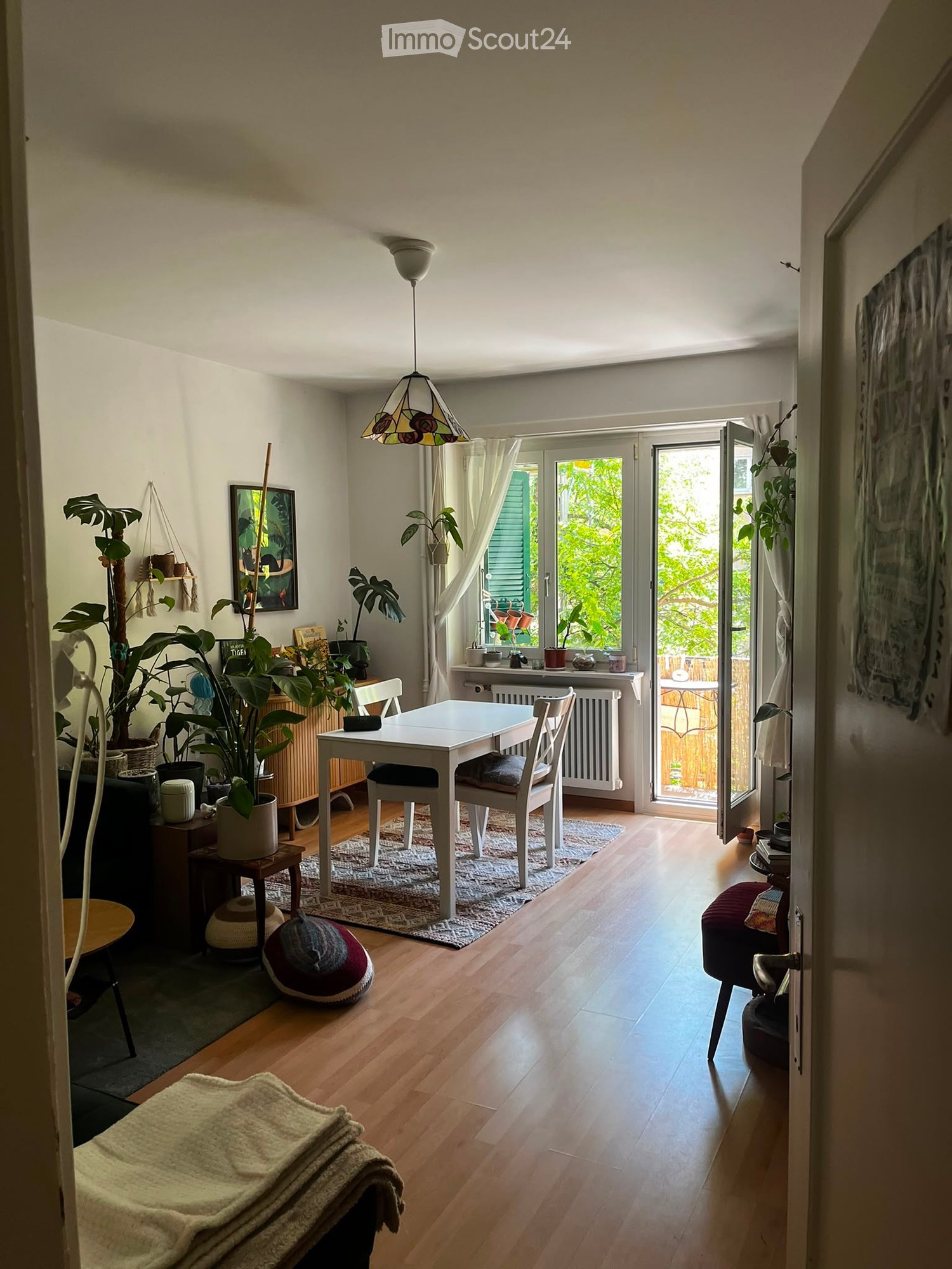 Bright living and dining area with wooden flooring, large windows, and a balcony. Features a white dining table with chairs, a sofa, a stool, and several potted plants.