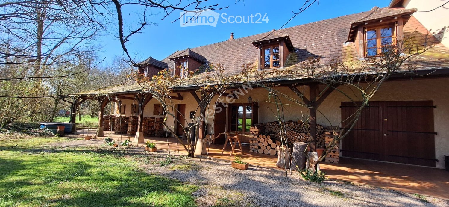 A two-story house with a tiled roof, wooden beams, and a covered porch area. The house features a mix of stone and wood exterior, with a well-maintained garden and stacks of firewood nearby.