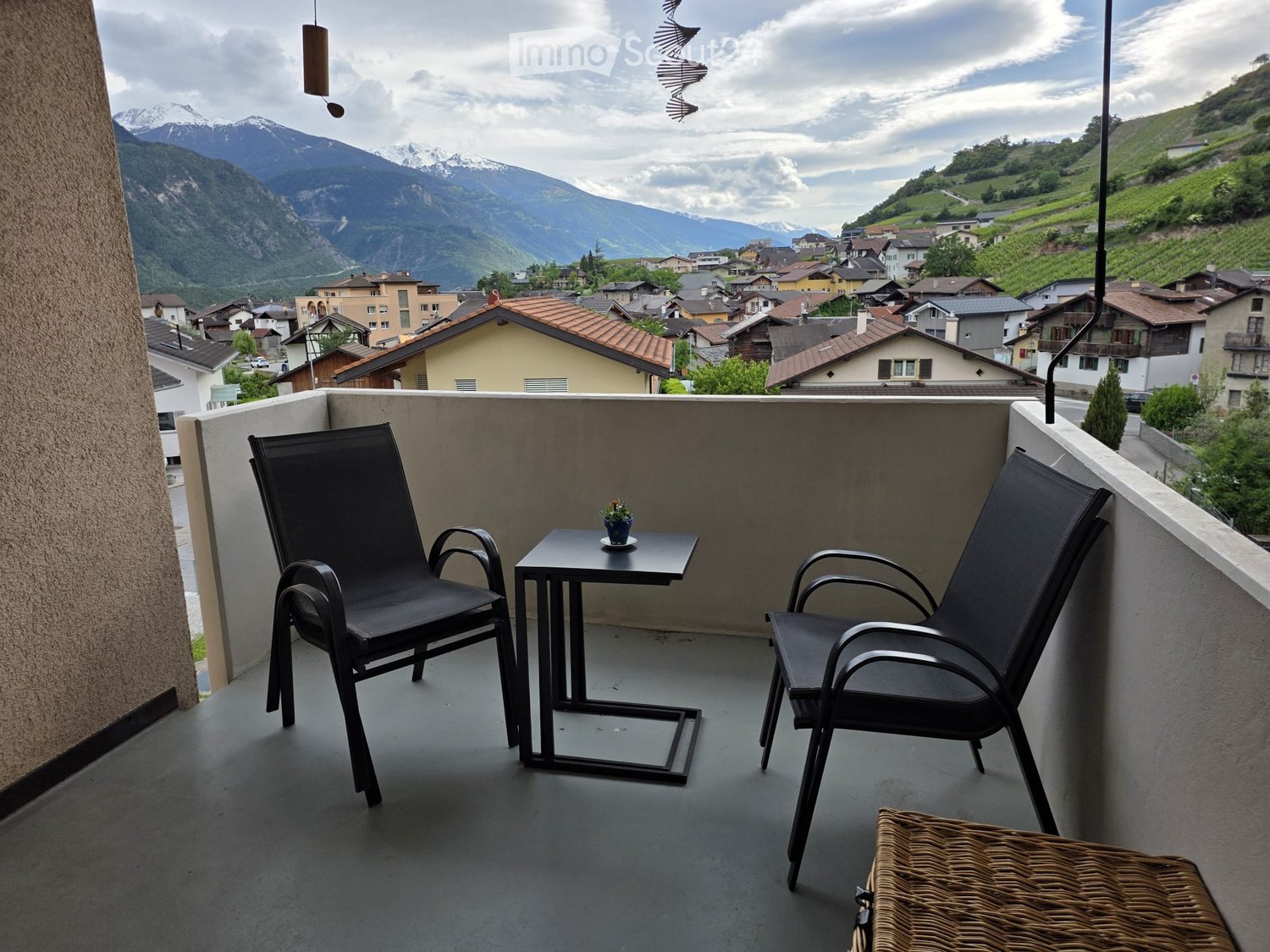 Balcony with two black chairs, a small table, and a wicker chair, overlooking a scenic mountain town.