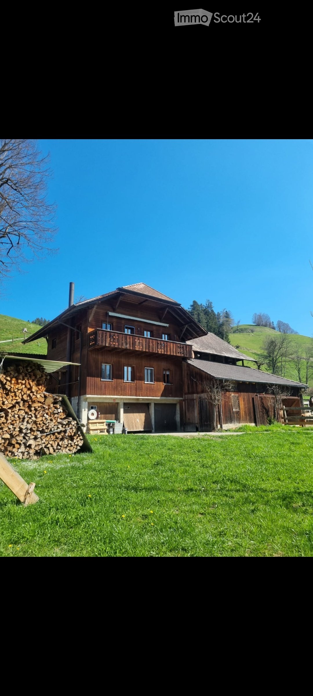 A two-story wooden house with a balcony, surrounded by a grassy lawn and a stack of firewood.