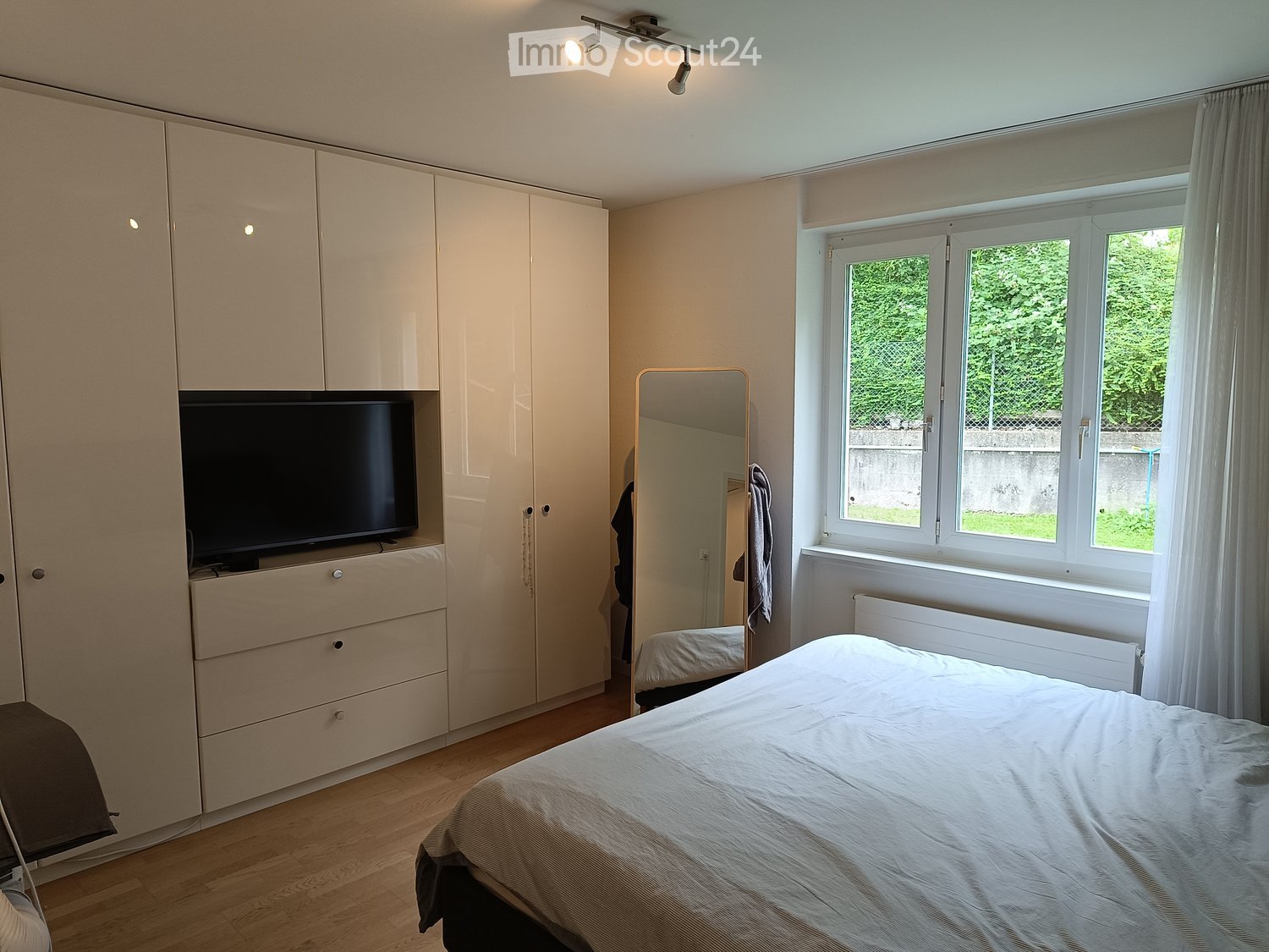 Modern bedroom with a large bed, built-in wardrobe, TV, mirror, and large window with curtains.