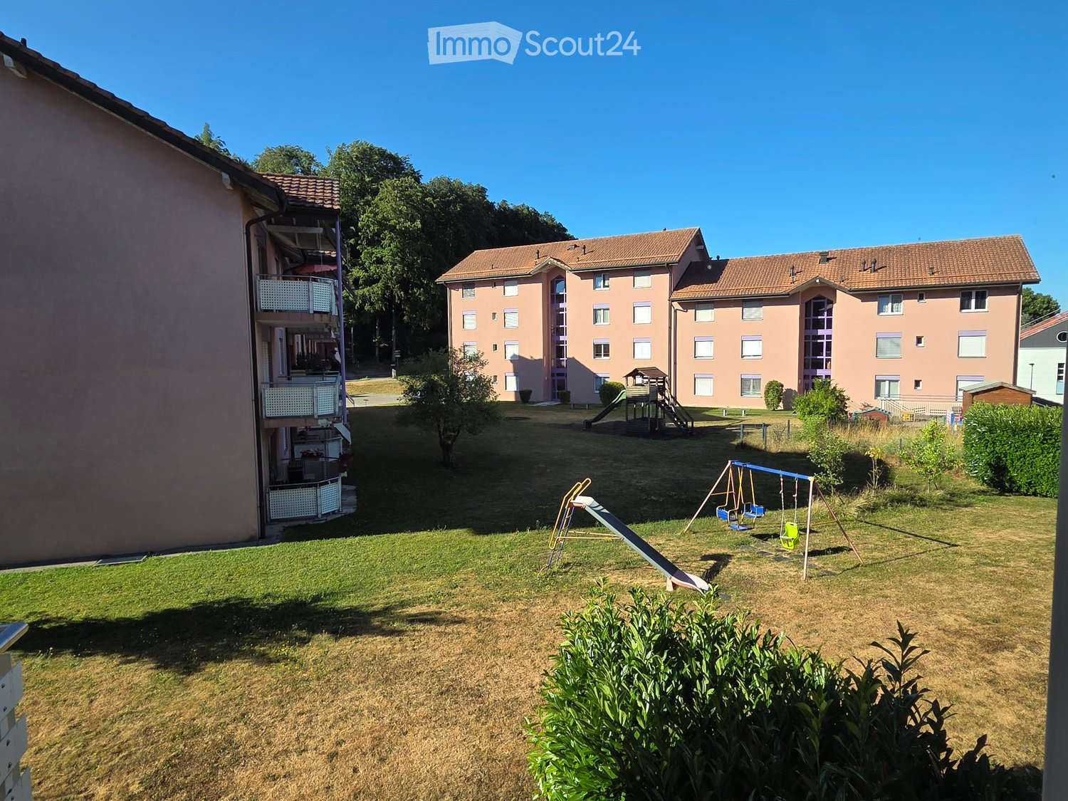 Residential complex with a grassy courtyard, playground equipment including a slide and swings, and surrounding buildings with balconies and windows.