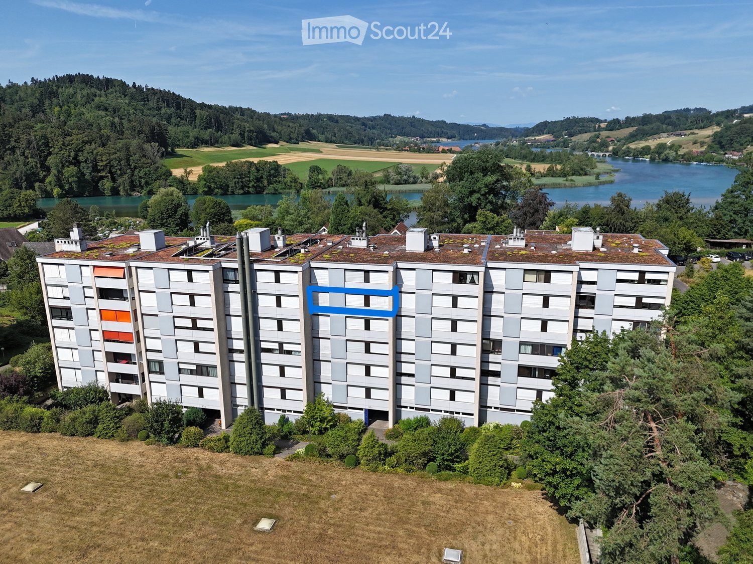 Multi-story residential building with grey facade, multiple windows, and a flat roof with greenery, surrounded by trees and a body of water in the background.