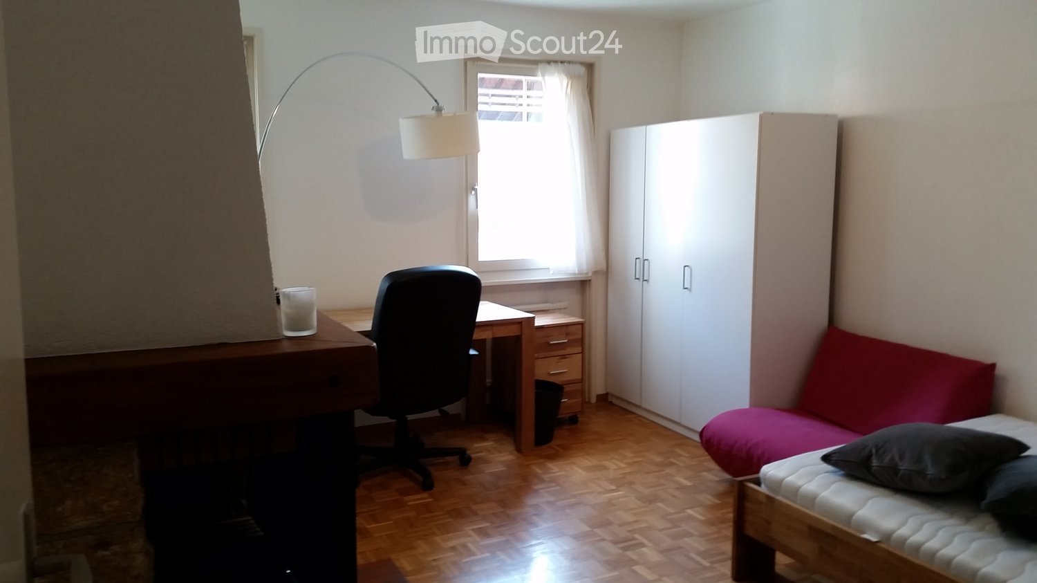 A small, well-lit room with a wooden floor, a white wall-mounted wardrobe, a red sofa, a desk with a chair, and a floor lamp near the window.