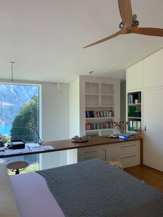 Modern bedroom with a large window offering a scenic mountain view, a bed with a grey duvet, a wooden desk with a chair, a bookshelf filled with books, and a ceiling fan.