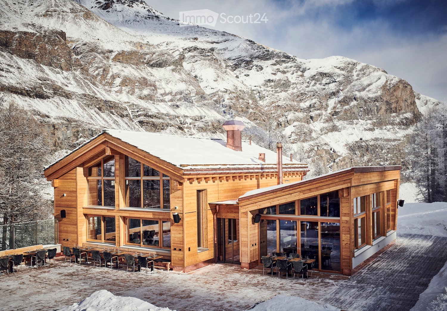 A large wooden building with snow-covered roof and large windows, situated in a snowy mountain landscape. The building has an outdoor seating area with tables and chairs.