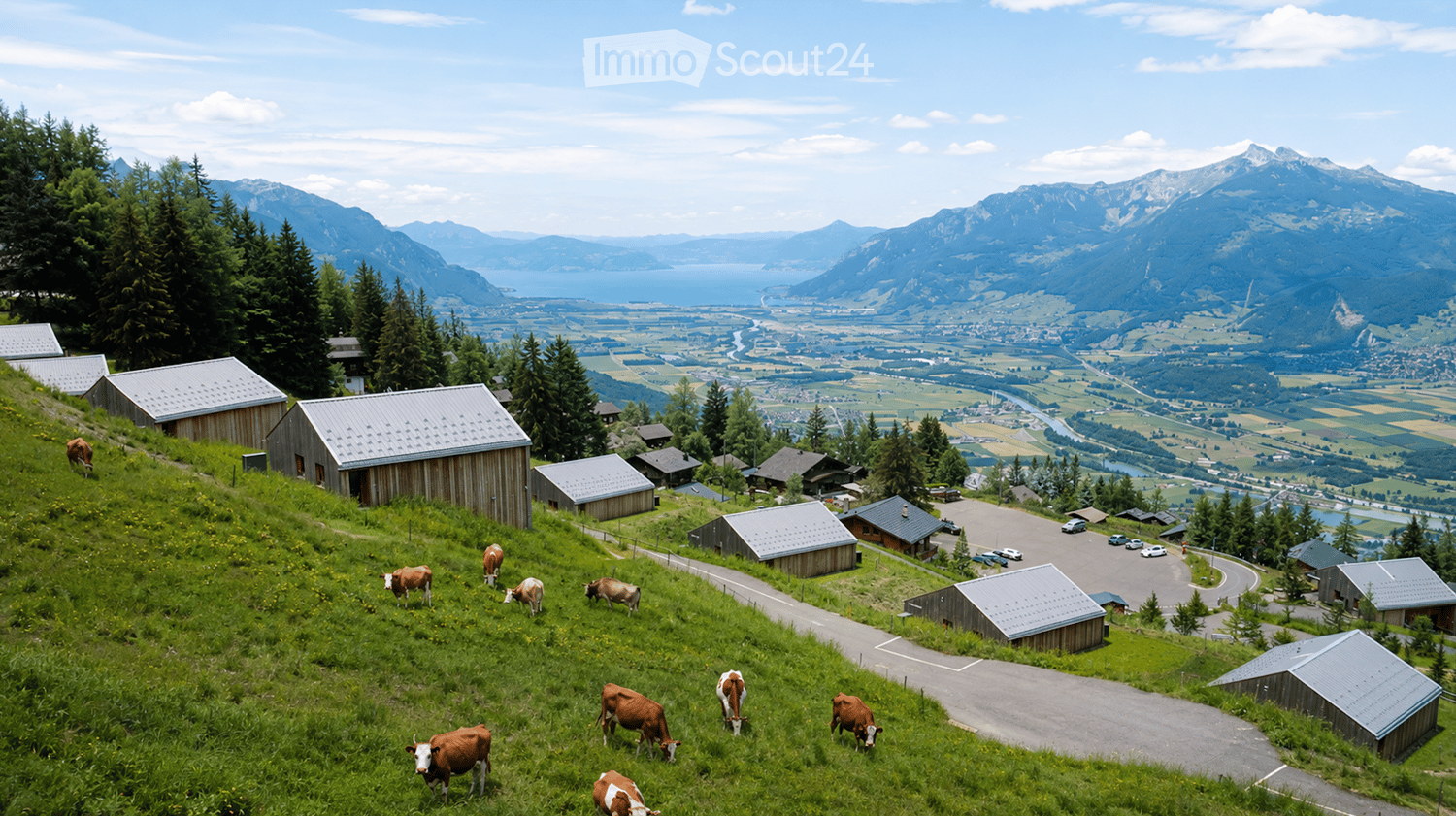 A scenic view of a rural area with cows grazing on a green hillside, surrounded by wooden barns and a winding road, with a vast valley and mountain range in the background.