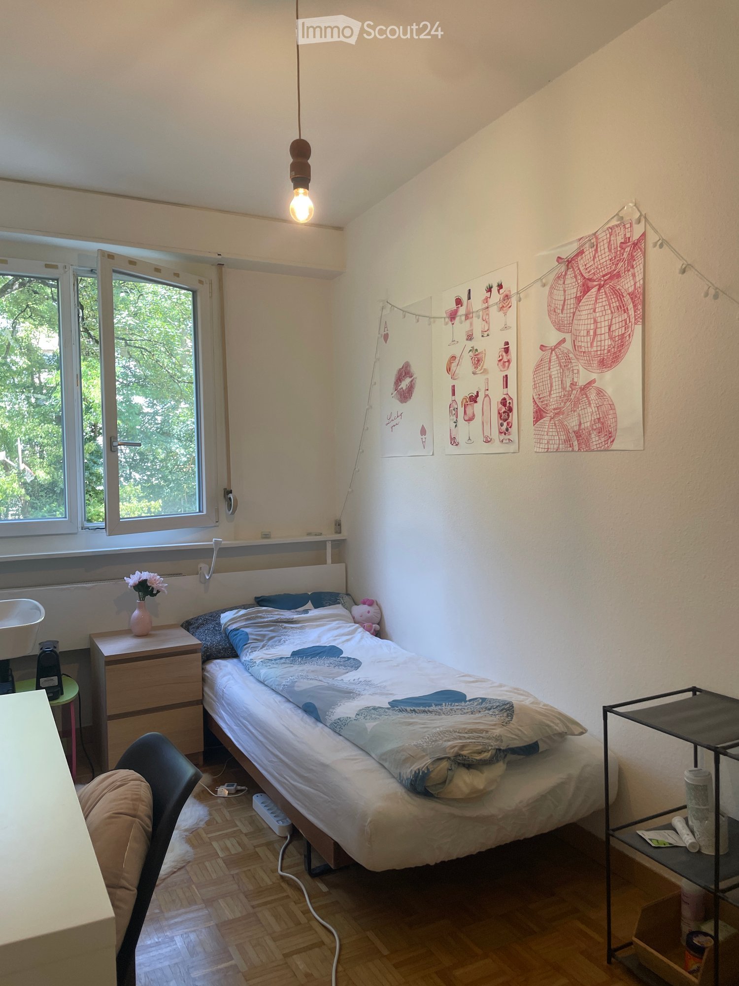 Cozy bedroom with a single bed, nightstand, chair, and a small table. Features wooden flooring, large windows with a view of greenery, and decorative artwork on the walls.