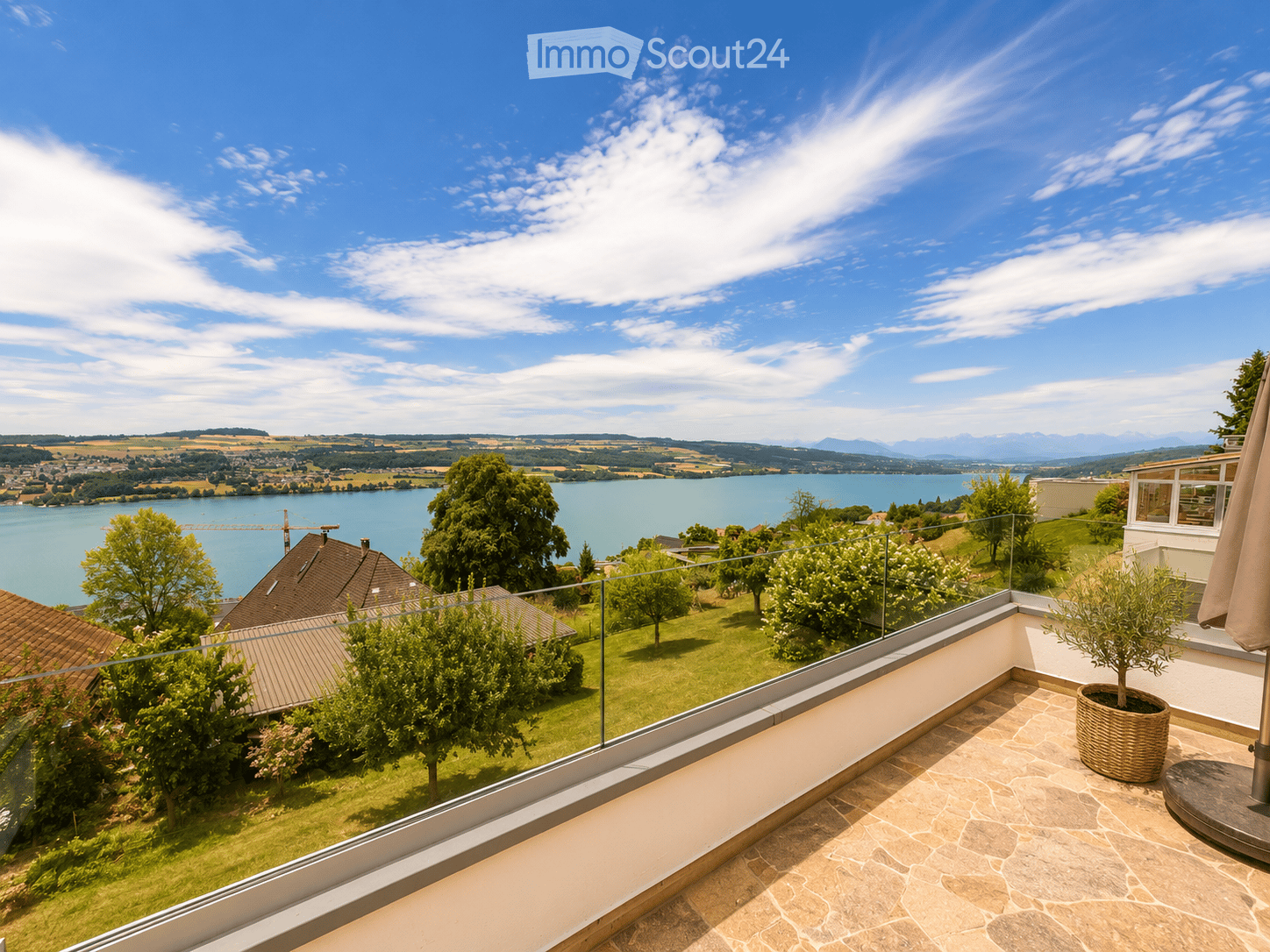 Balcony with a view of a lake and surrounding landscape, featuring a table, umbrella, and potted plant.