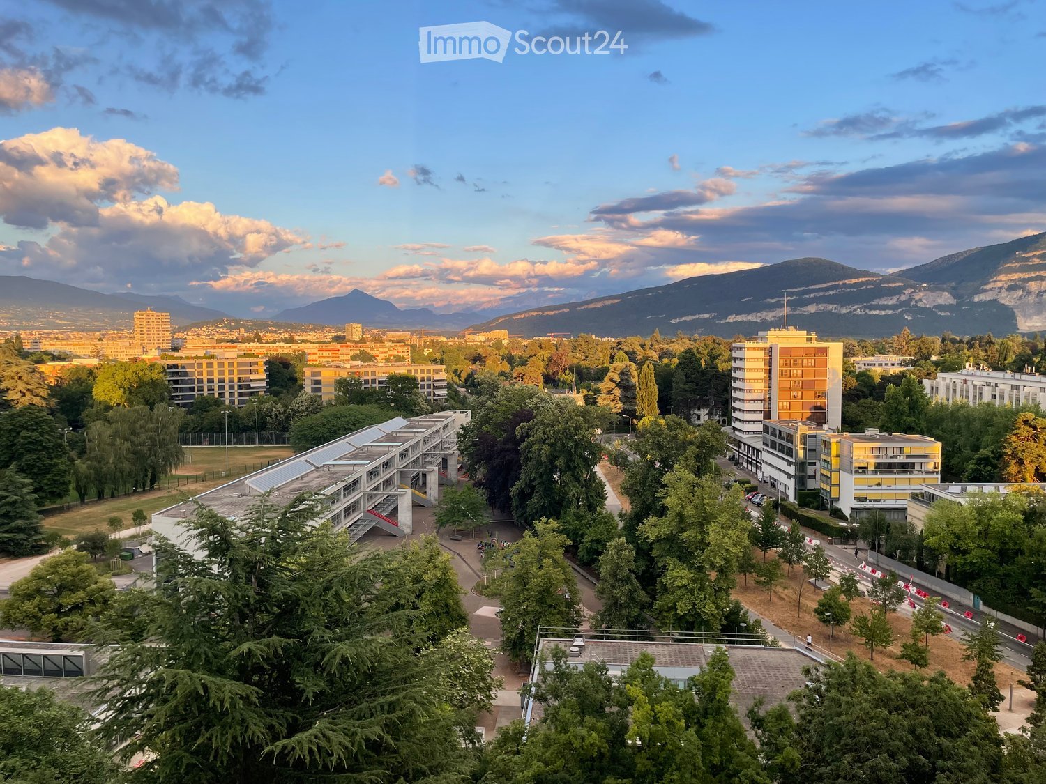Aerial view of a university campus with multiple buildings, trees, and a distant mountain range under a partly cloudy sky.