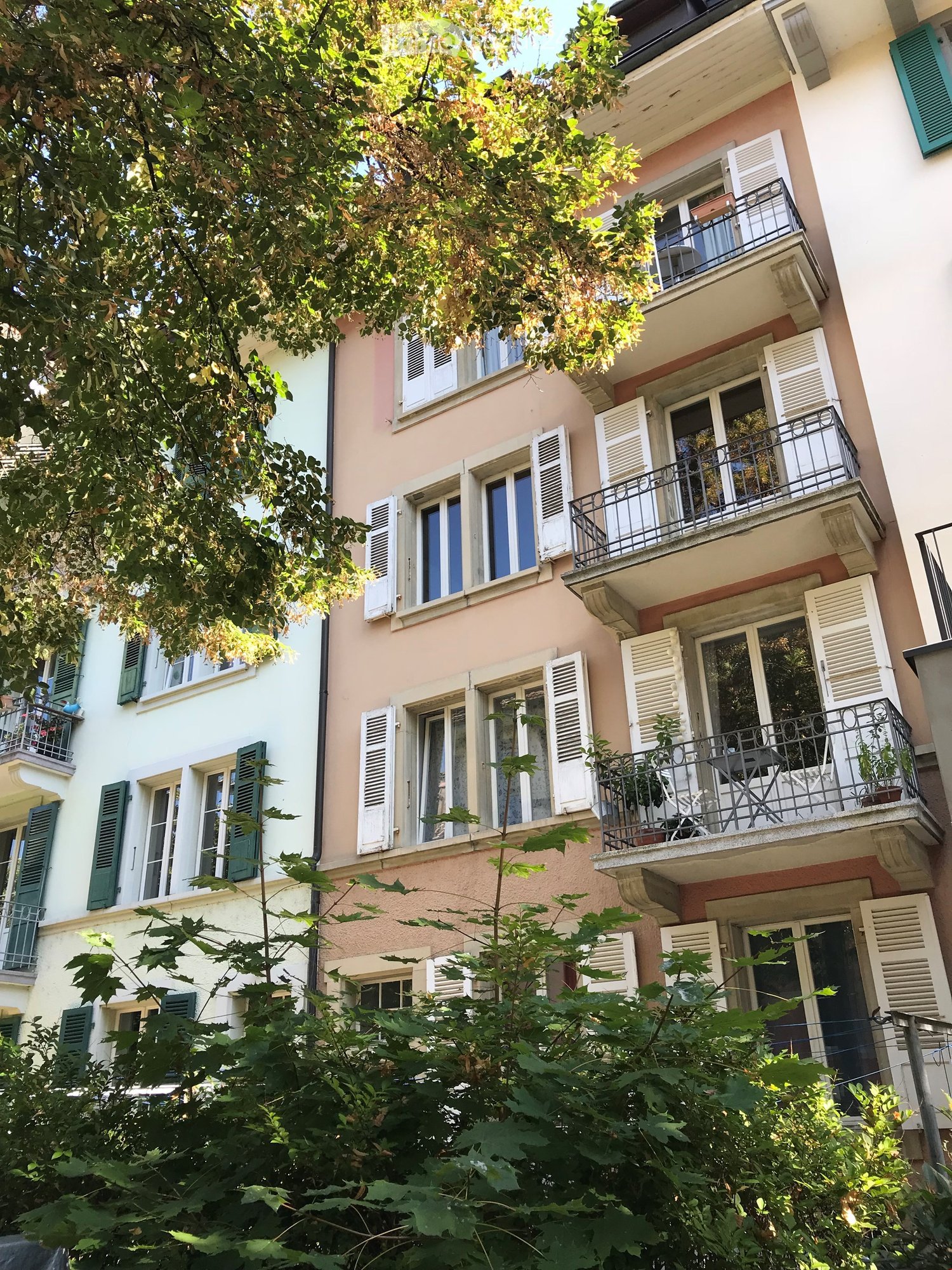 A multi-story residential building with light pink and white facades, multiple windows with shutters, and balconies adorned with plants and outdoor furniture.