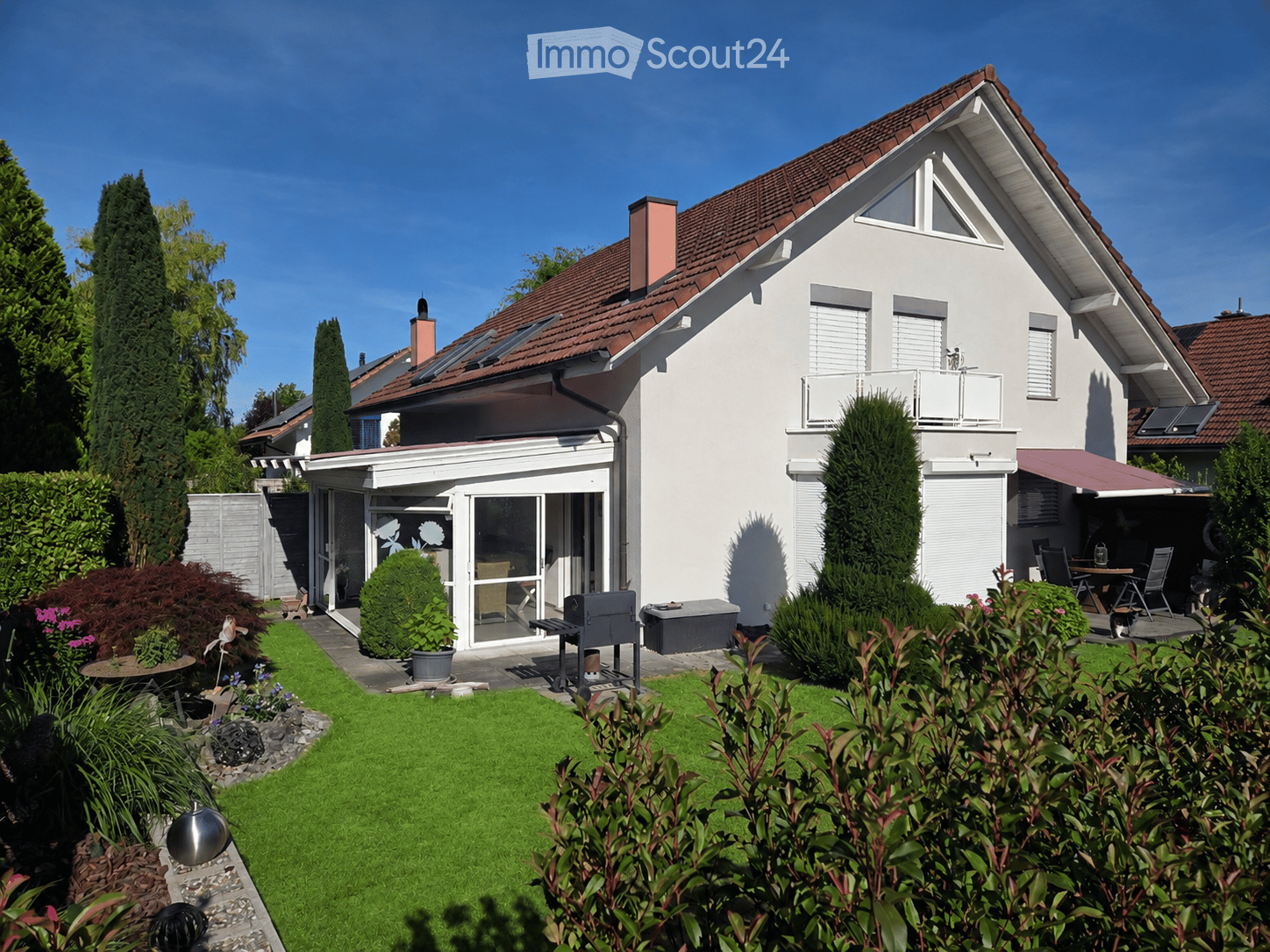 Two-story house with white exterior, red-tiled roof, and a well-maintained garden featuring a lawn, shrubs, and a patio area with outdoor furniture.
