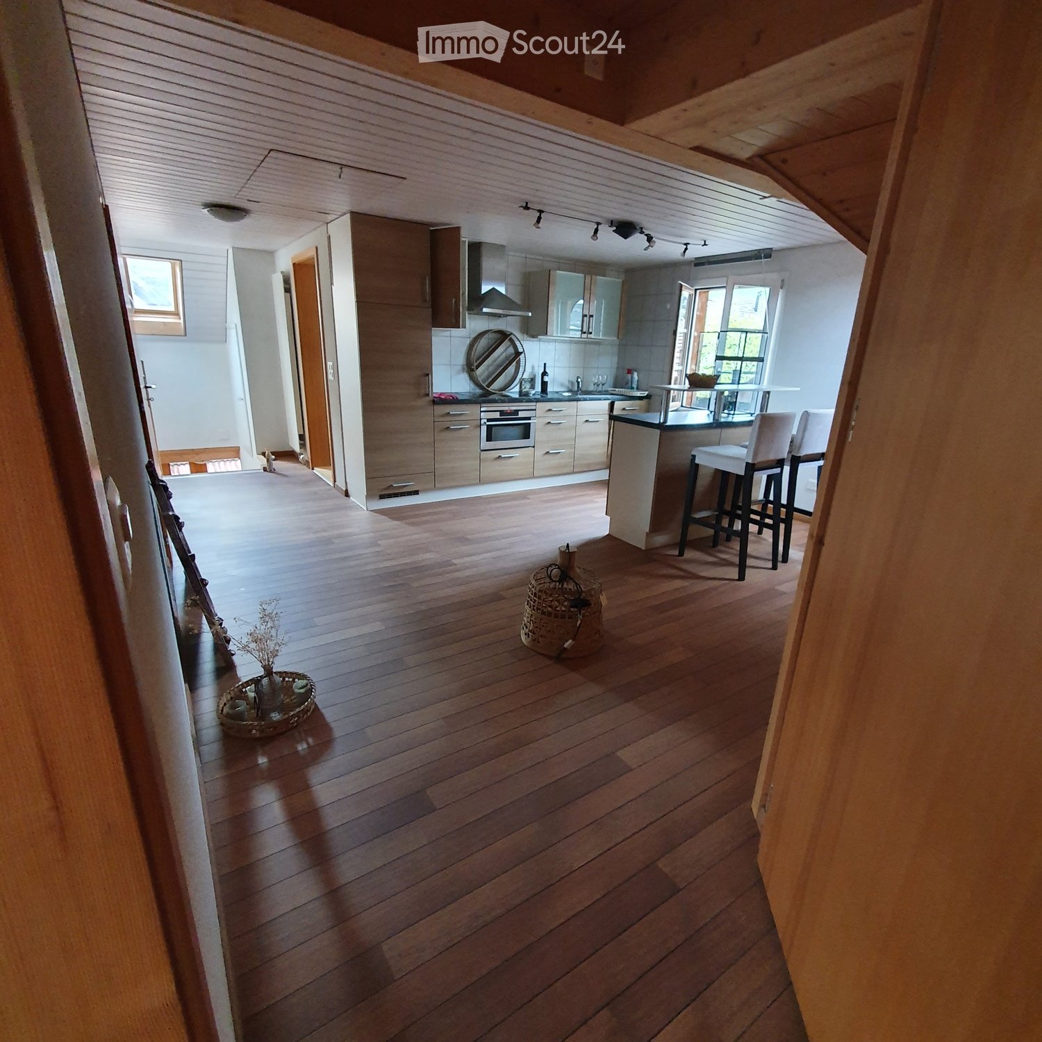 Open-plan living and kitchen area with wooden flooring, modern kitchen cabinets, and bar stools.